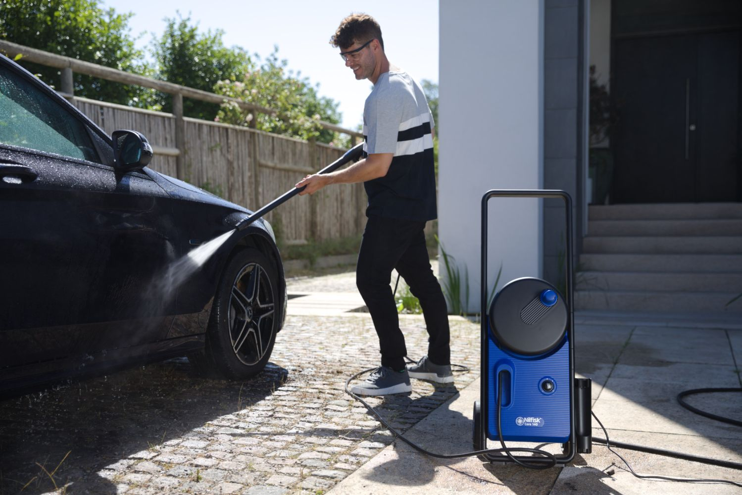 Uomo lava auto nera con idropulitrice davanti a casa moderna.