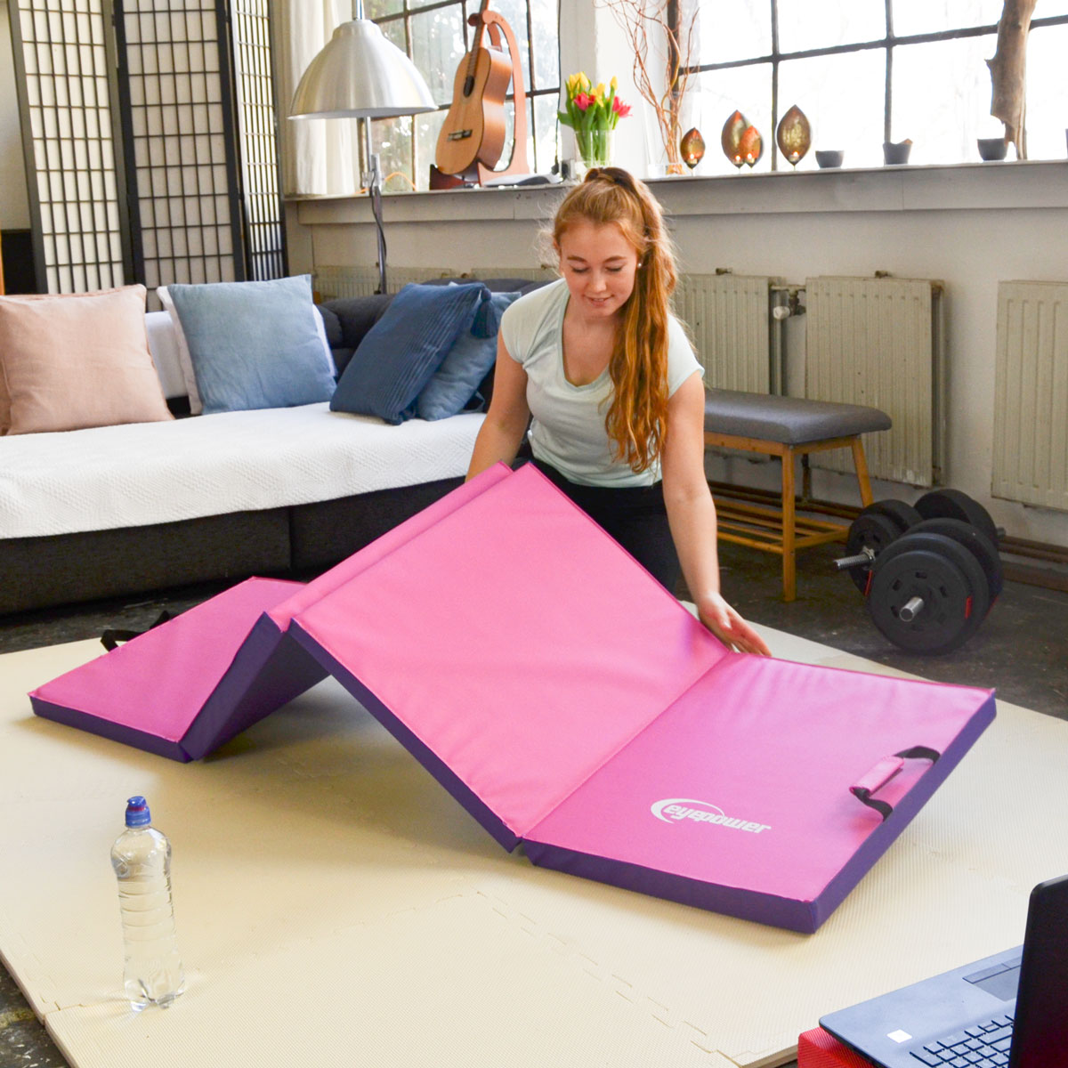 Eine Frau breitet eine pinke Matte aus. Hanteln und ein Sofa sind im Hintergrund.