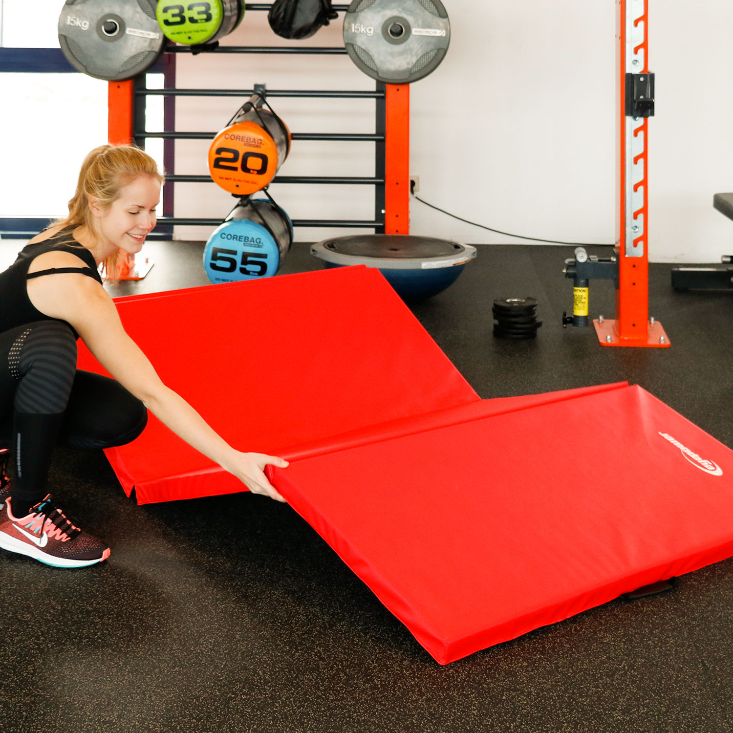 Eine Frau legt eine rote Matte in einem Fitnessstudio aus. Im Hintergrund sind Gewichte und Ausrüstung.