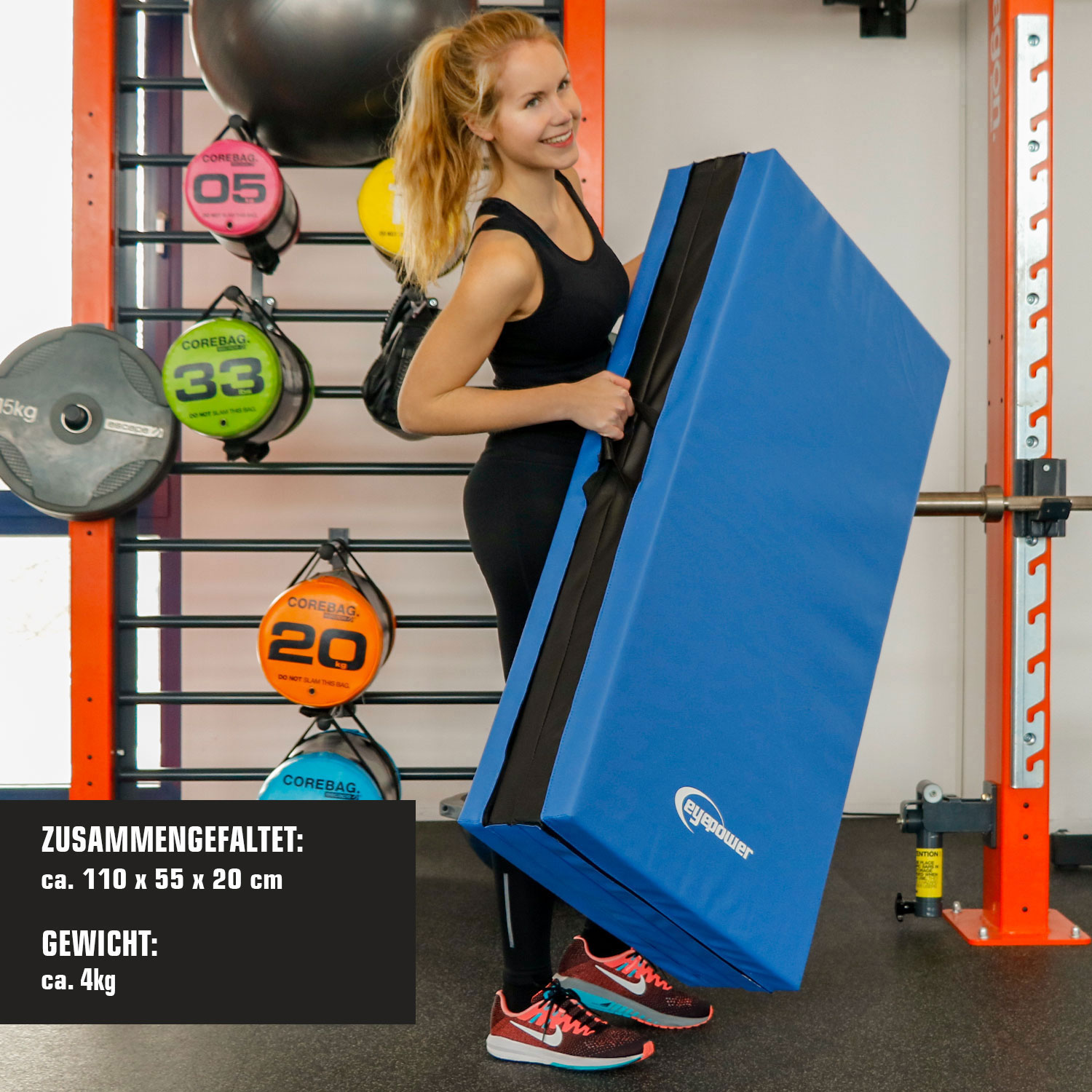 Frau hält eine blaue Matte in einem Fitnessstudio mit Trainingsgeräten im Hintergrund.
