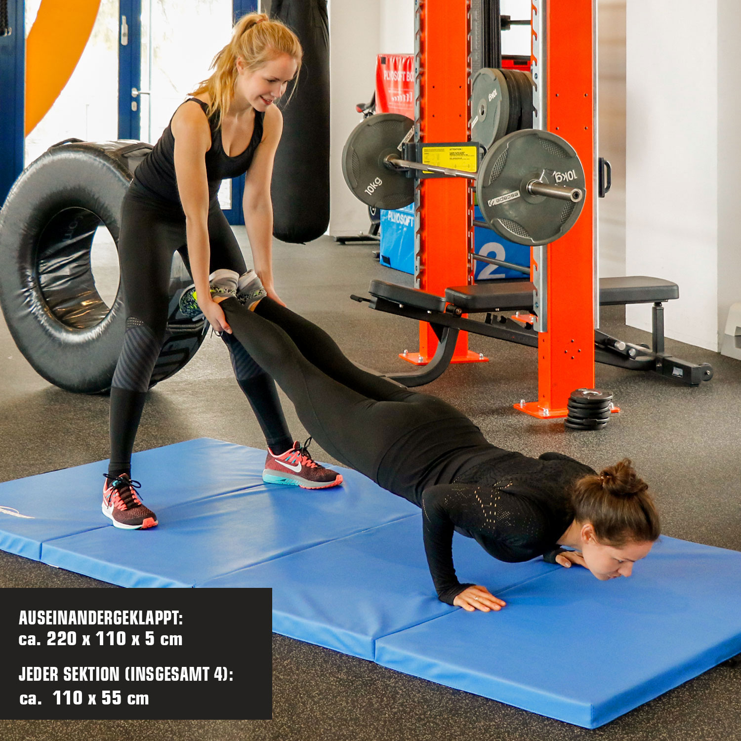 Zwei Frauen trainieren in einem Fitnessstudio, eine macht Liegestütze auf einer blauen Matte, die andere hilft.