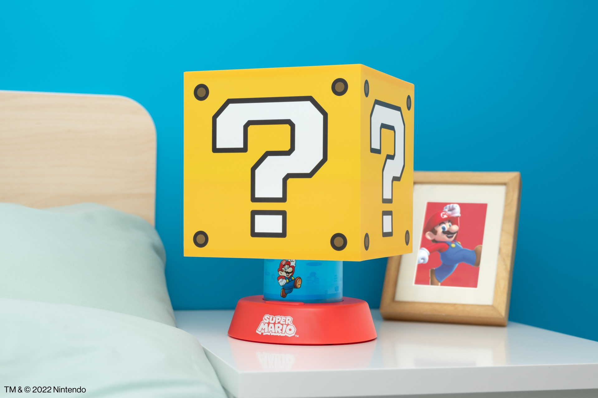 Żółta lampa Super Mario w kształcie bloku z pytajnikiem na szafce nocnej, obok łóżka i obrazka.