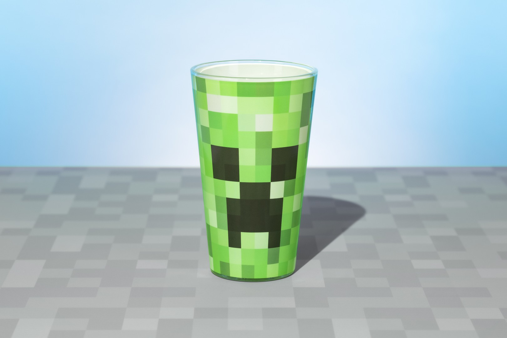Zielony kubek Minecraft Creeper z pikselową twarzą na szarej kratownicy.