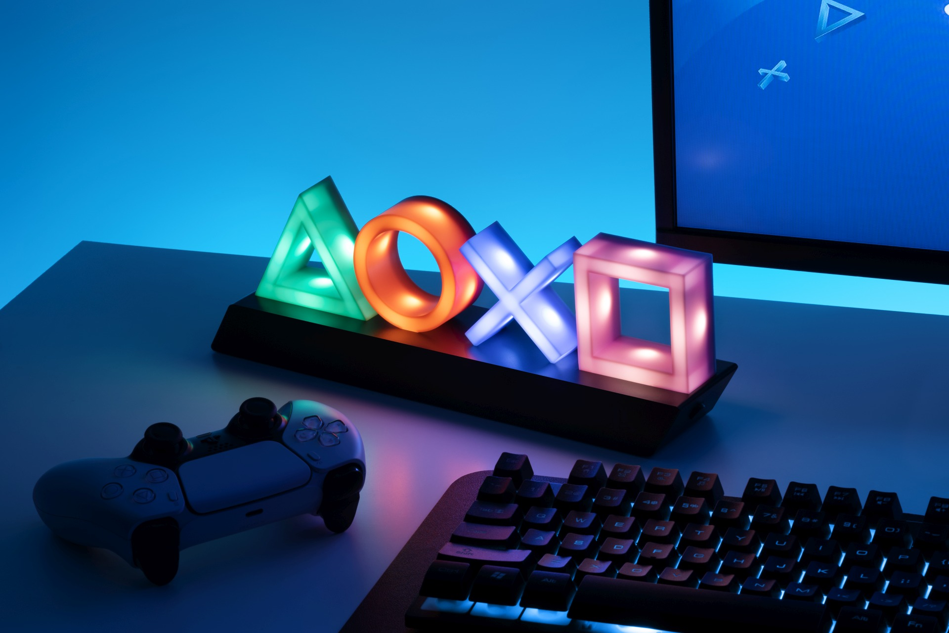 Leuchtende PlayStation-Symbole (Dreieck, Kreis, Kreuz, Quadrat) auf einem Schreibtisch mit Controller und Tastatur.