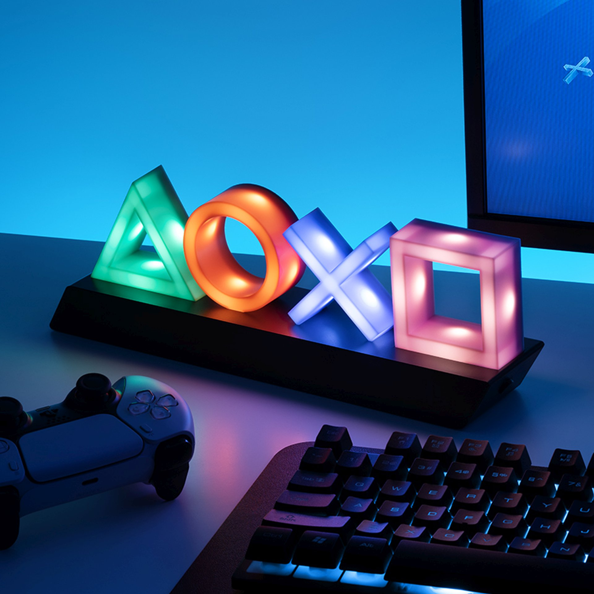 Leuchtende PlayStation-Symbole auf einem Schreibtisch mit Controller und Tastatur.