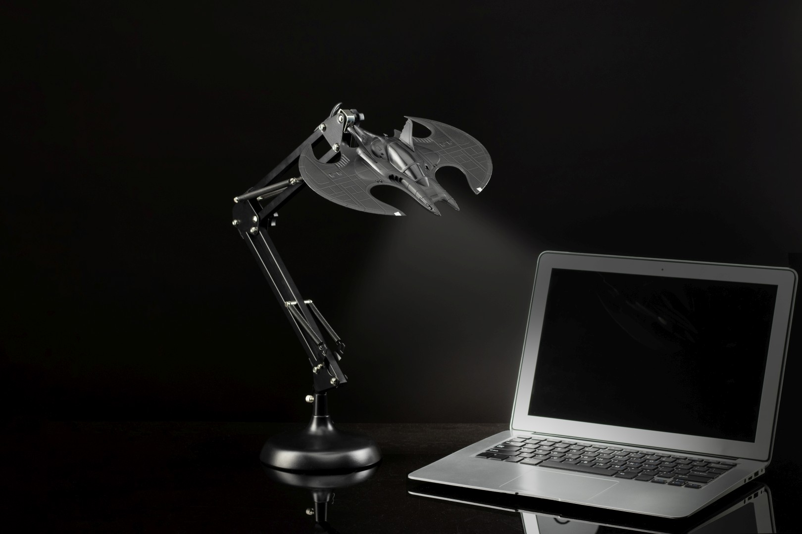 Eine schwarze, fledermausförmige Lampe beleuchtet einen silbernen Laptop auf einer dunklen, reflektierenden Oberfläche.
