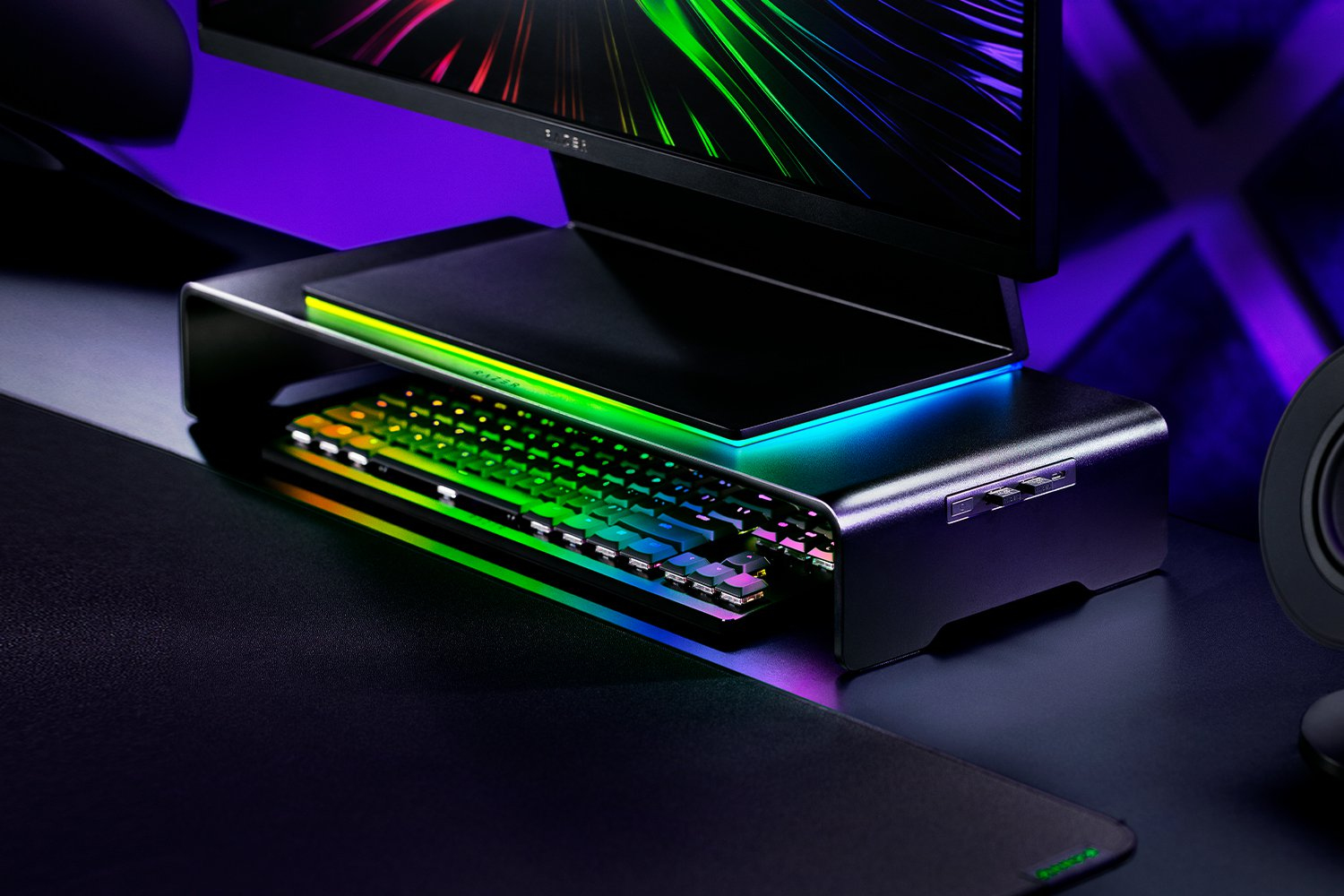 Eine Gaming-Tastatur mit bunter RGB-Beleuchtung steht unter einem Monitorständer.