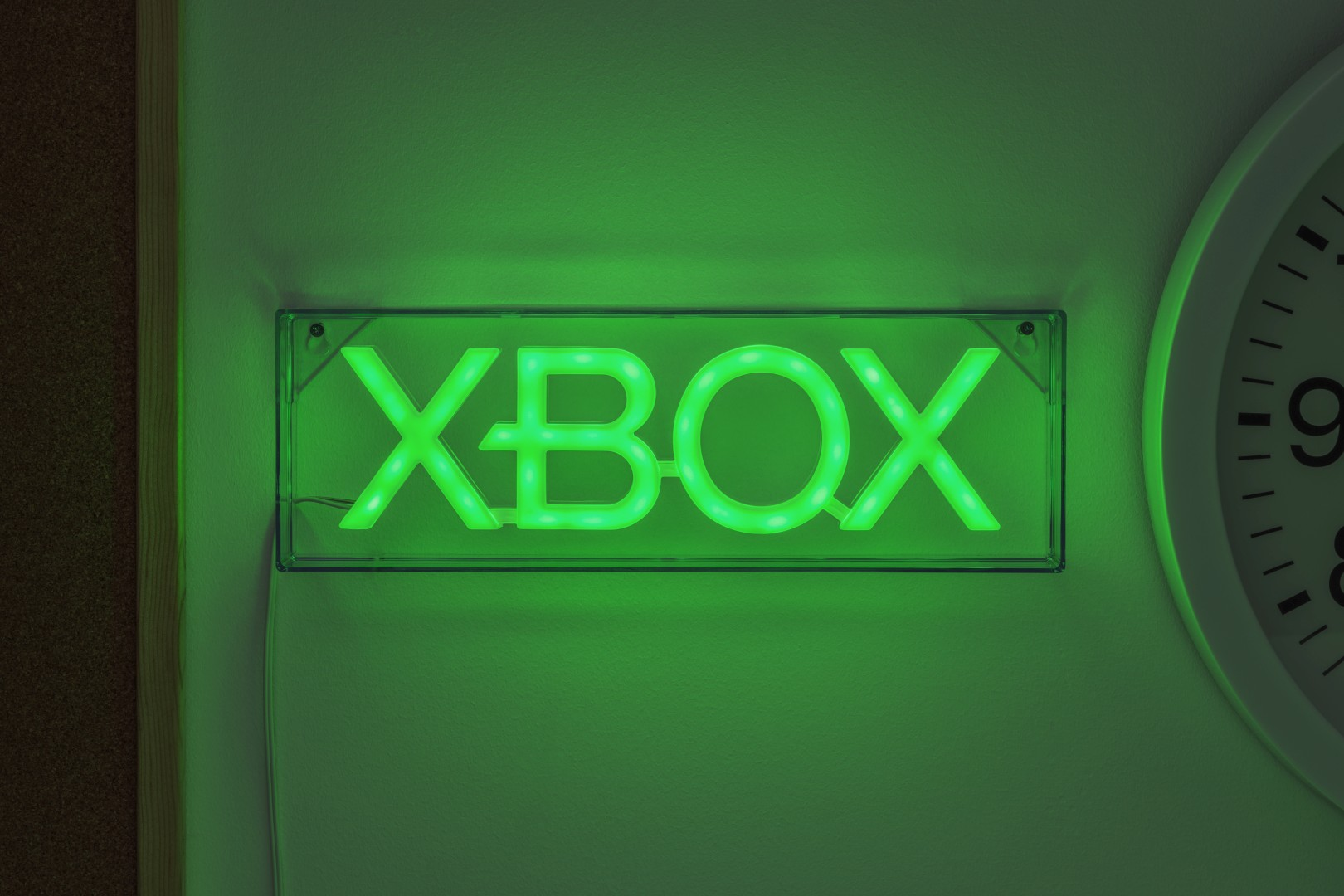 Zielony neonowy znak logo Xbox zamontowany na ścianie obok białego zegara.