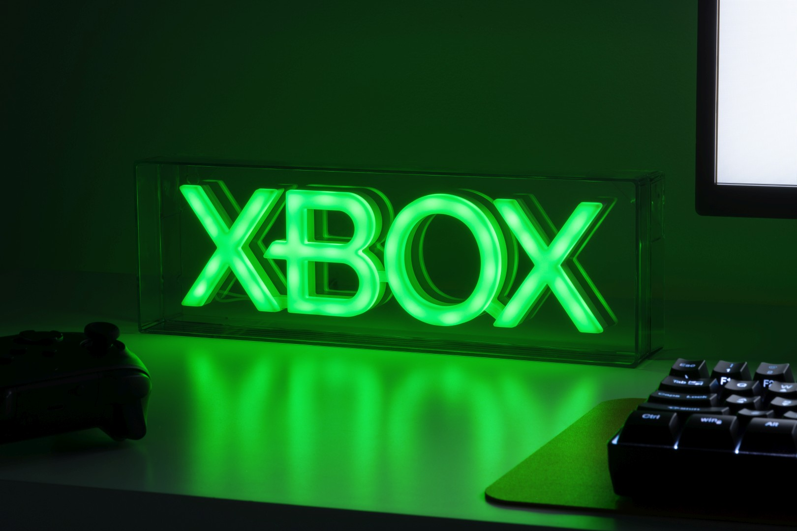 Zielone neonowe logo Xbox na biurku z kontrolerem i klawiaturą.
