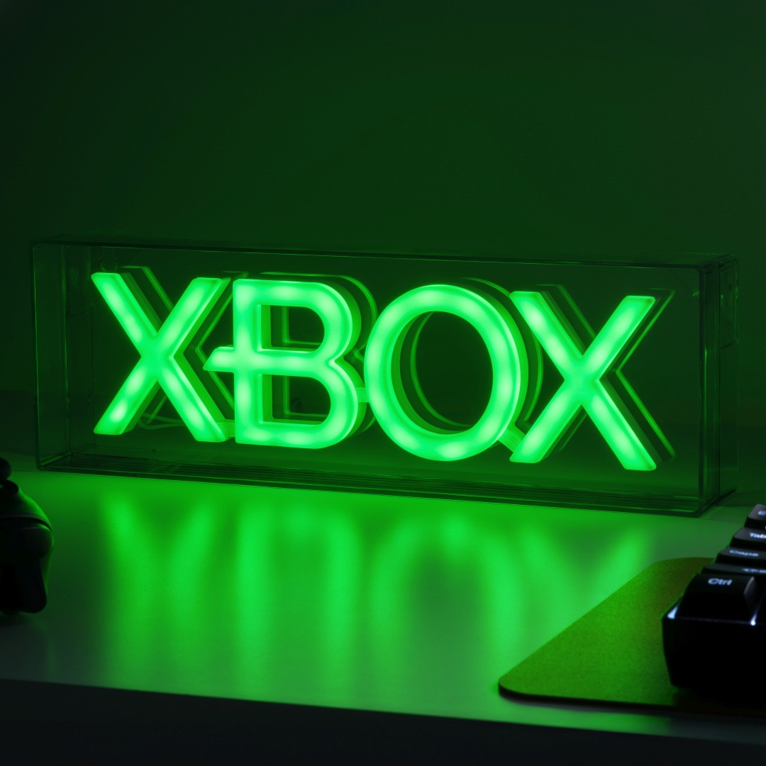 Zielone neonowe logo Xbox w przezroczystym pudełku, rzucające blask na biurko.