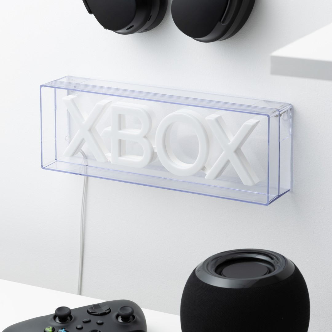 Białe litery Xbox w przezroczystym pudełku, obok kontroler i głośnik.