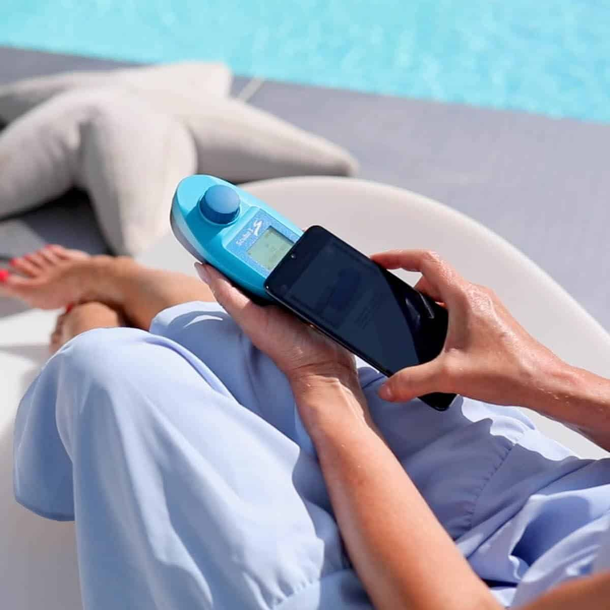 Una persona sostiene un medidor azul y un teléfono inteligente junto a una piscina.