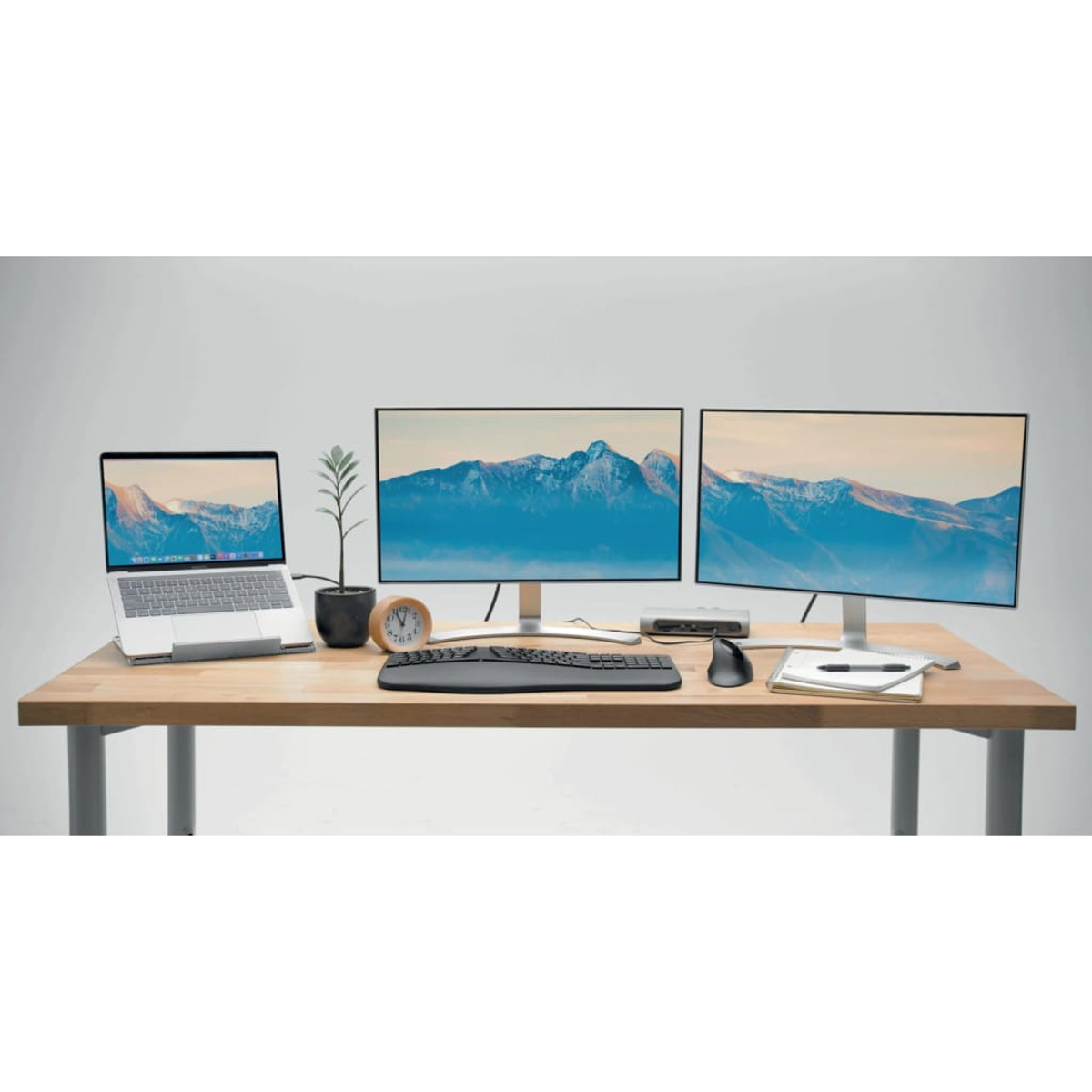 A modern desk setup with a laptop, two monitors, keyboard, mouse, and plant.