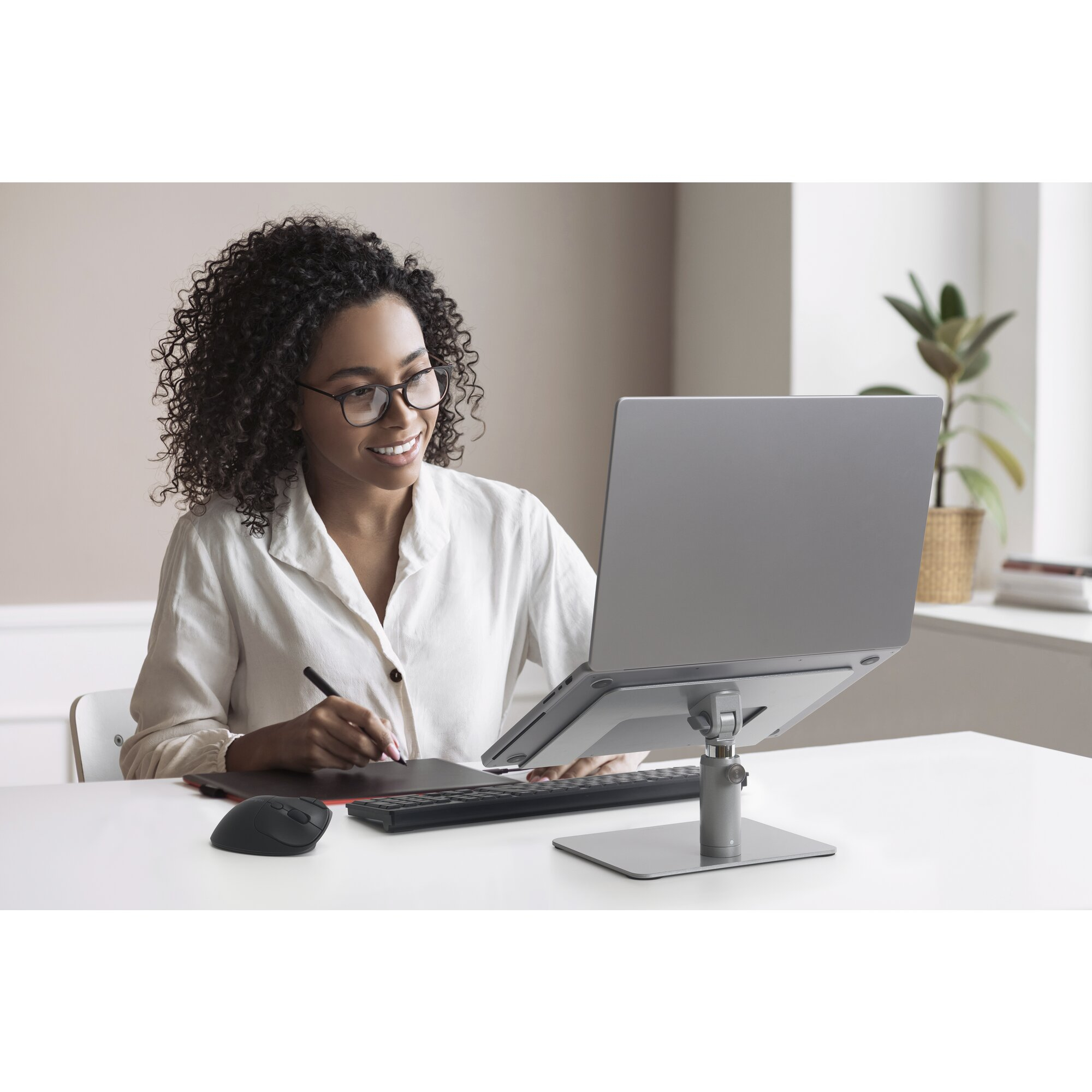 A woman with curly hair and glasses works on a laptop at a desk with a mouse and tablet.