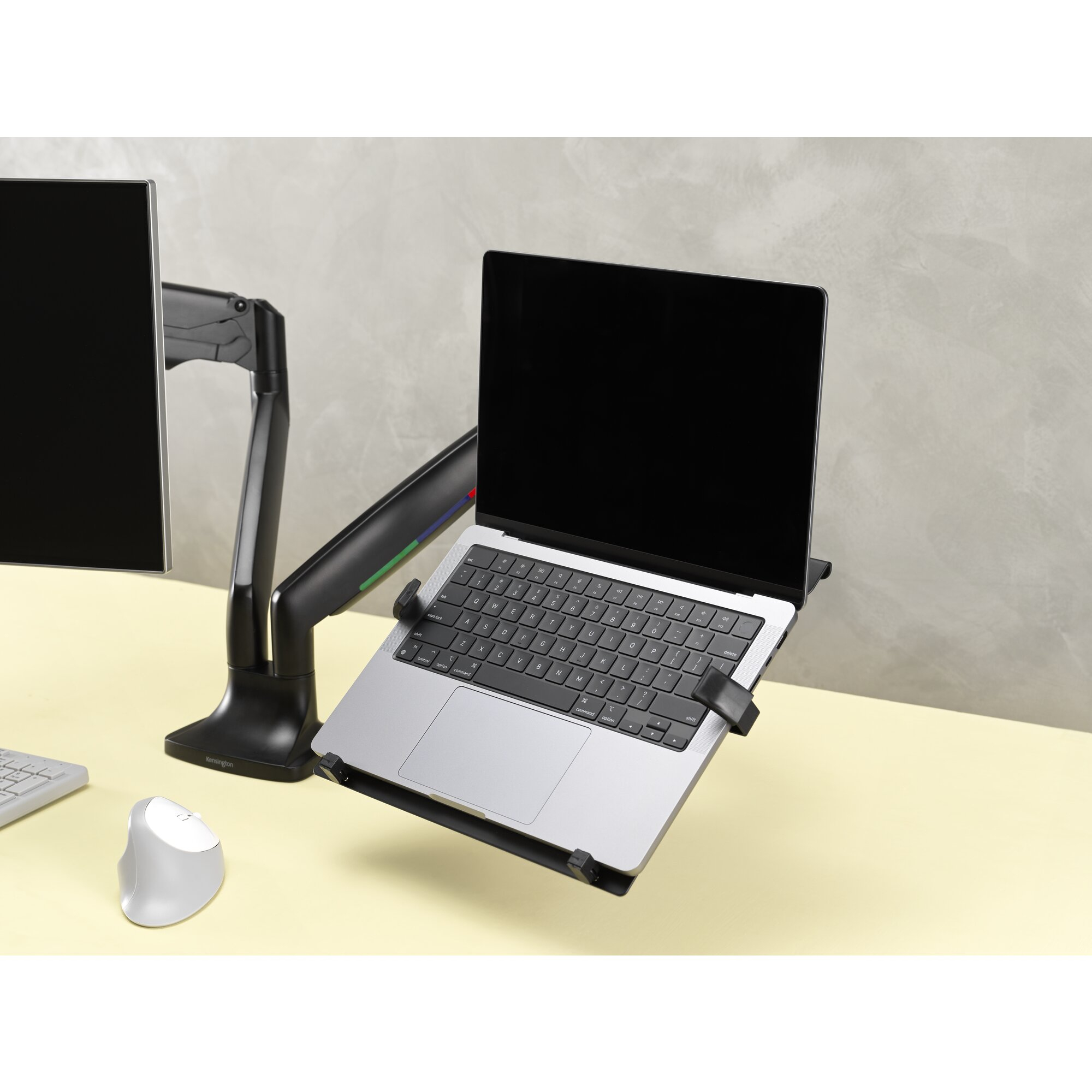 A silver laptop is mounted on a black arm, beside a black monitor. A white mouse is on the desk.
