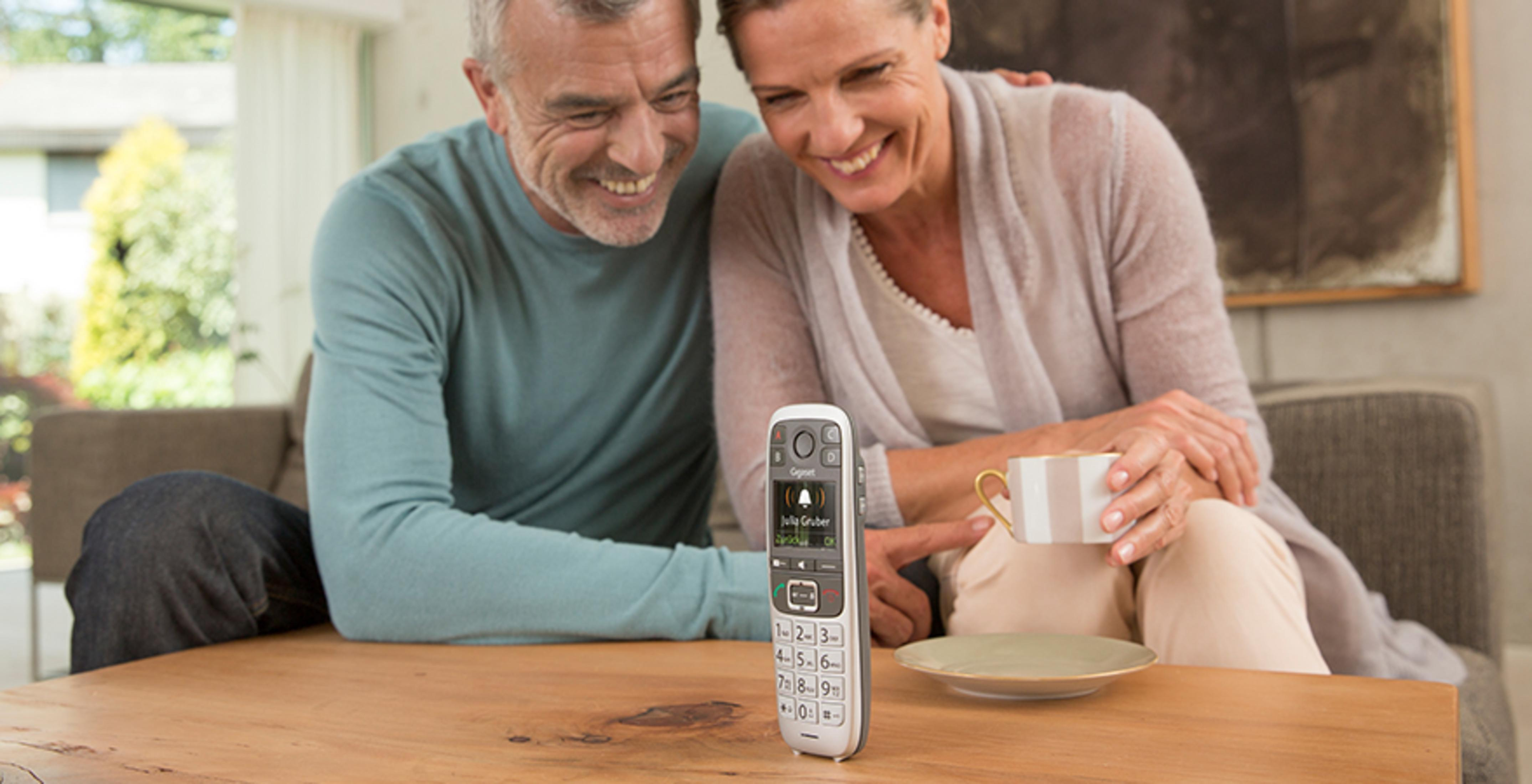 Una coppia anziana seduta a un tavolo di legno, sorridente. Un telefono al centro.