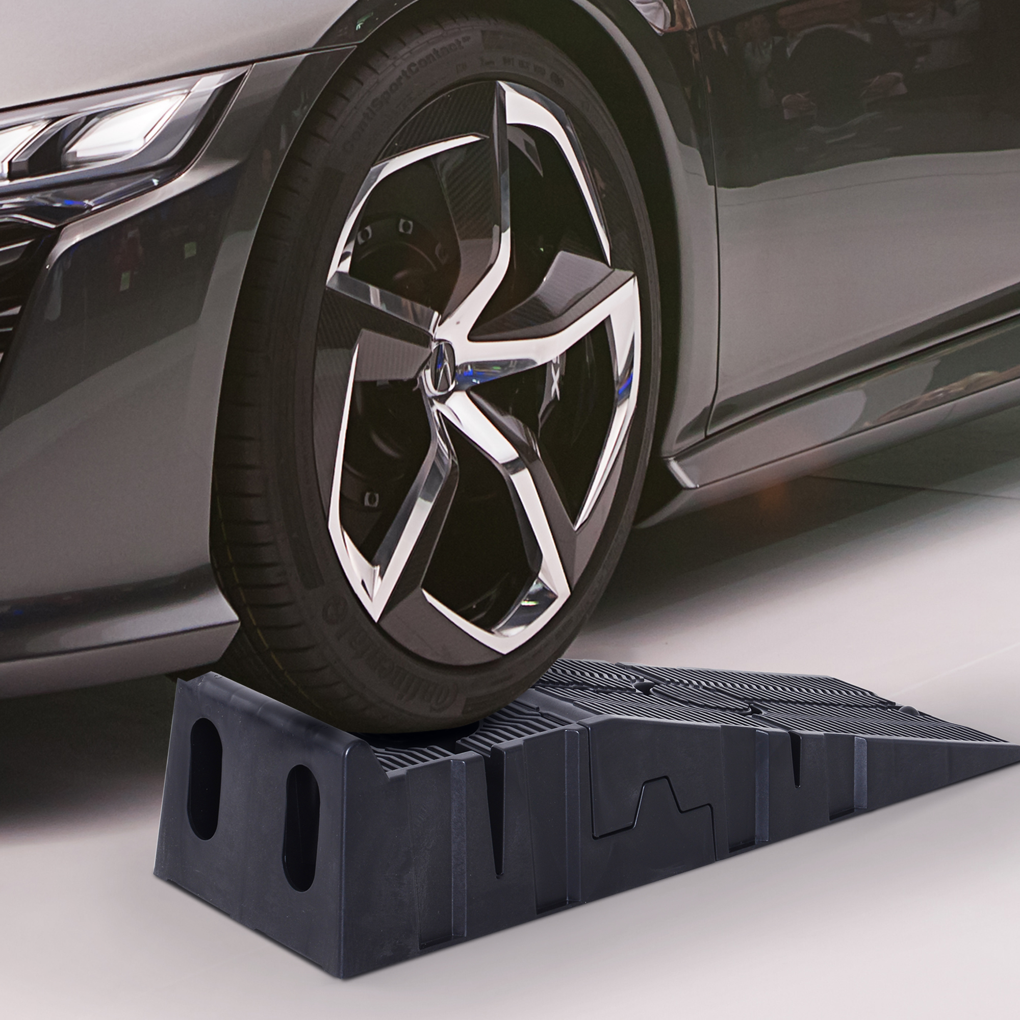 Un neumático de coche negro descansa sobre una rampa negra delante de un coche gris.