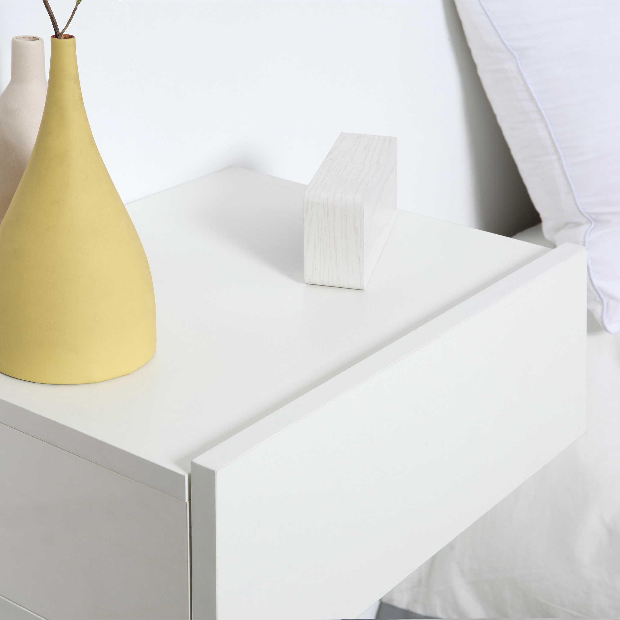 Un jarrón amarillo y uno beige sobre una mesita de noche blanca junto a una cama.
