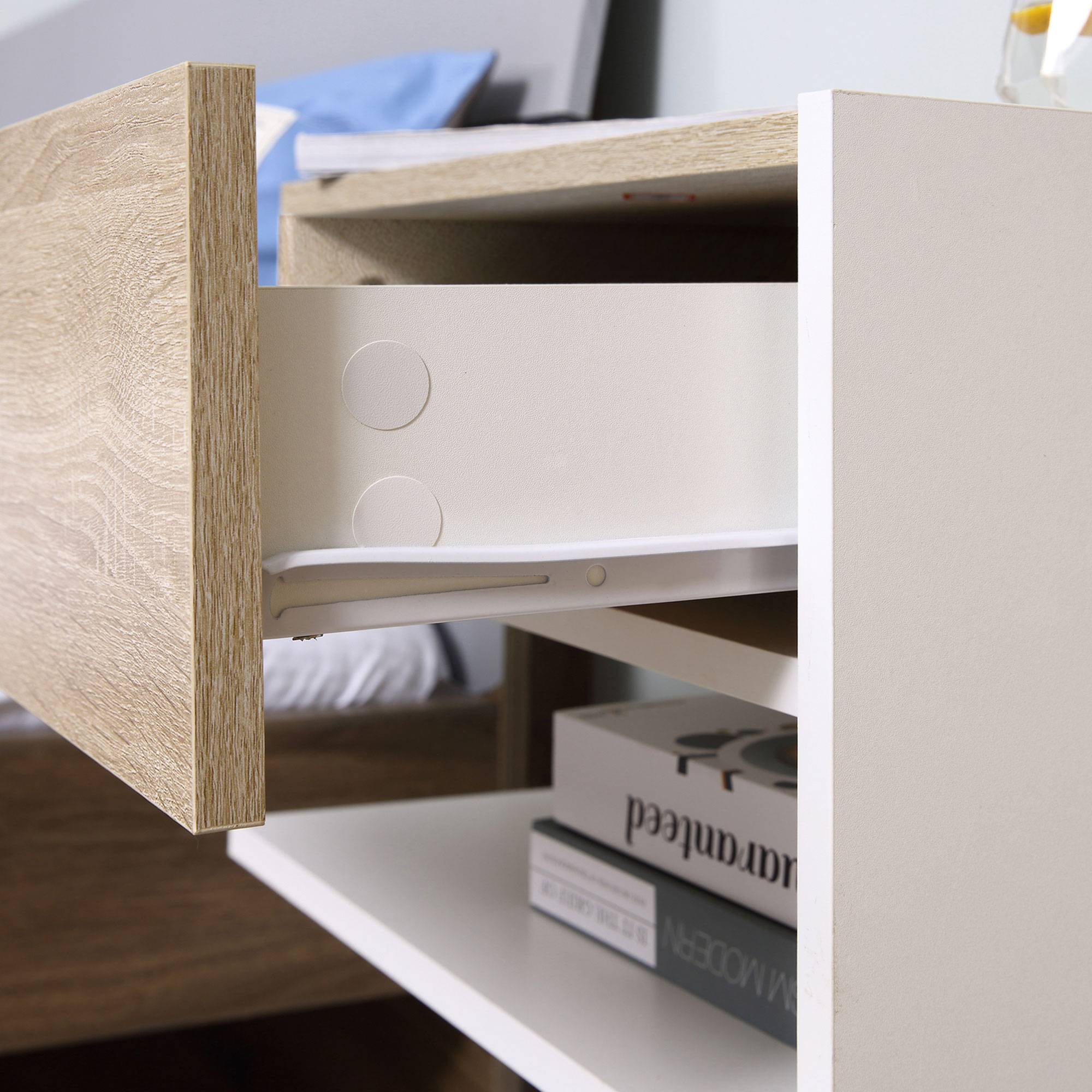 Mesita de noche con cajón abierto, acabado veteado en madera y cuerpo blanco. Libros en el estante.