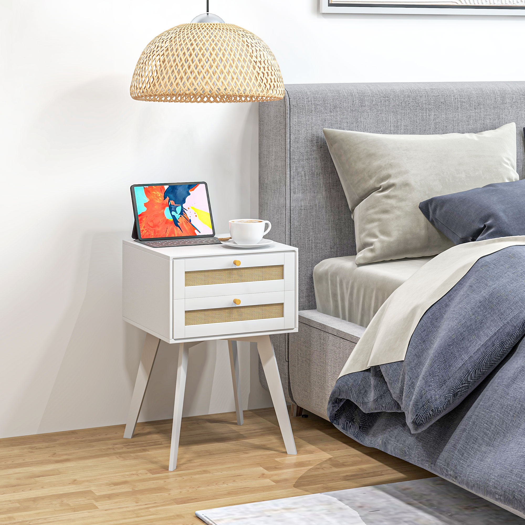 Una mesita de noche blanca con tableta, taza de café y detalle tejido junto a una cama.