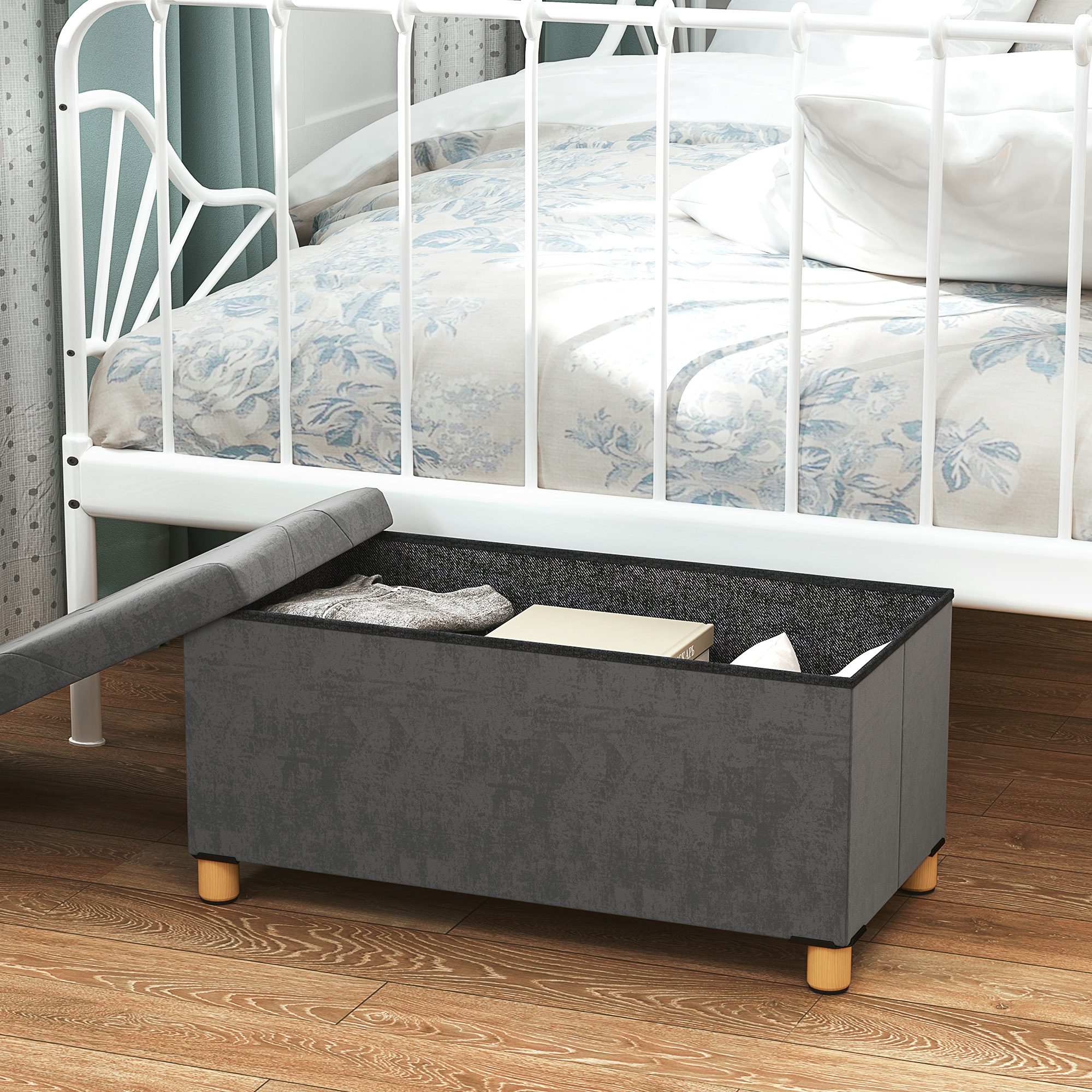 Banqueta de almacenamiento gris con tapa junto a una cama blanca con ropa de cama floral azul.