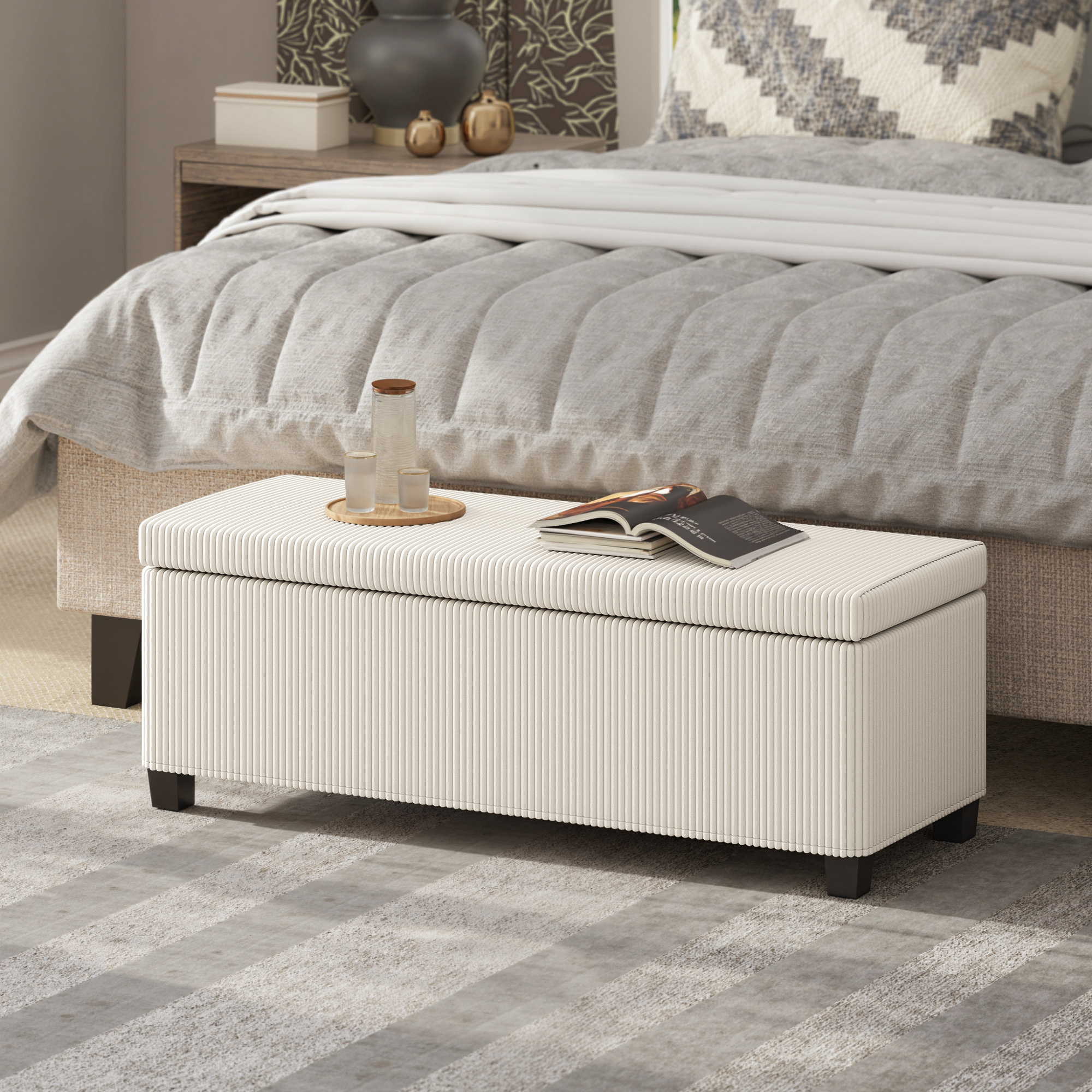 Un banco de almacenamiento beige se sienta frente a una cama con ropa de cama gris.
