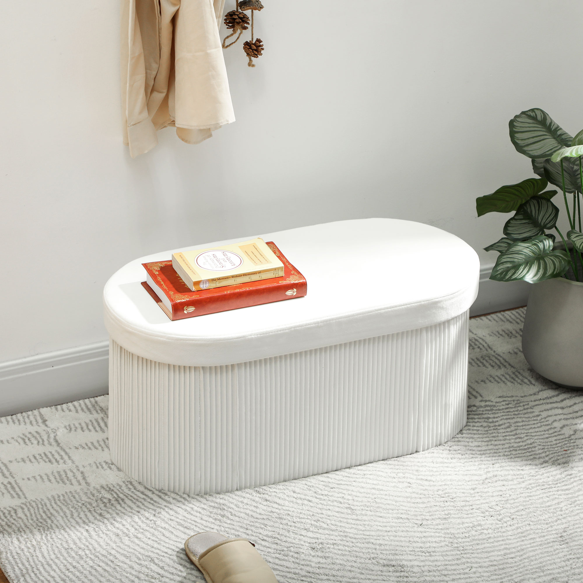 Otomana blanca con libros encima, abrigo beige y piñas colgadas en la pared.