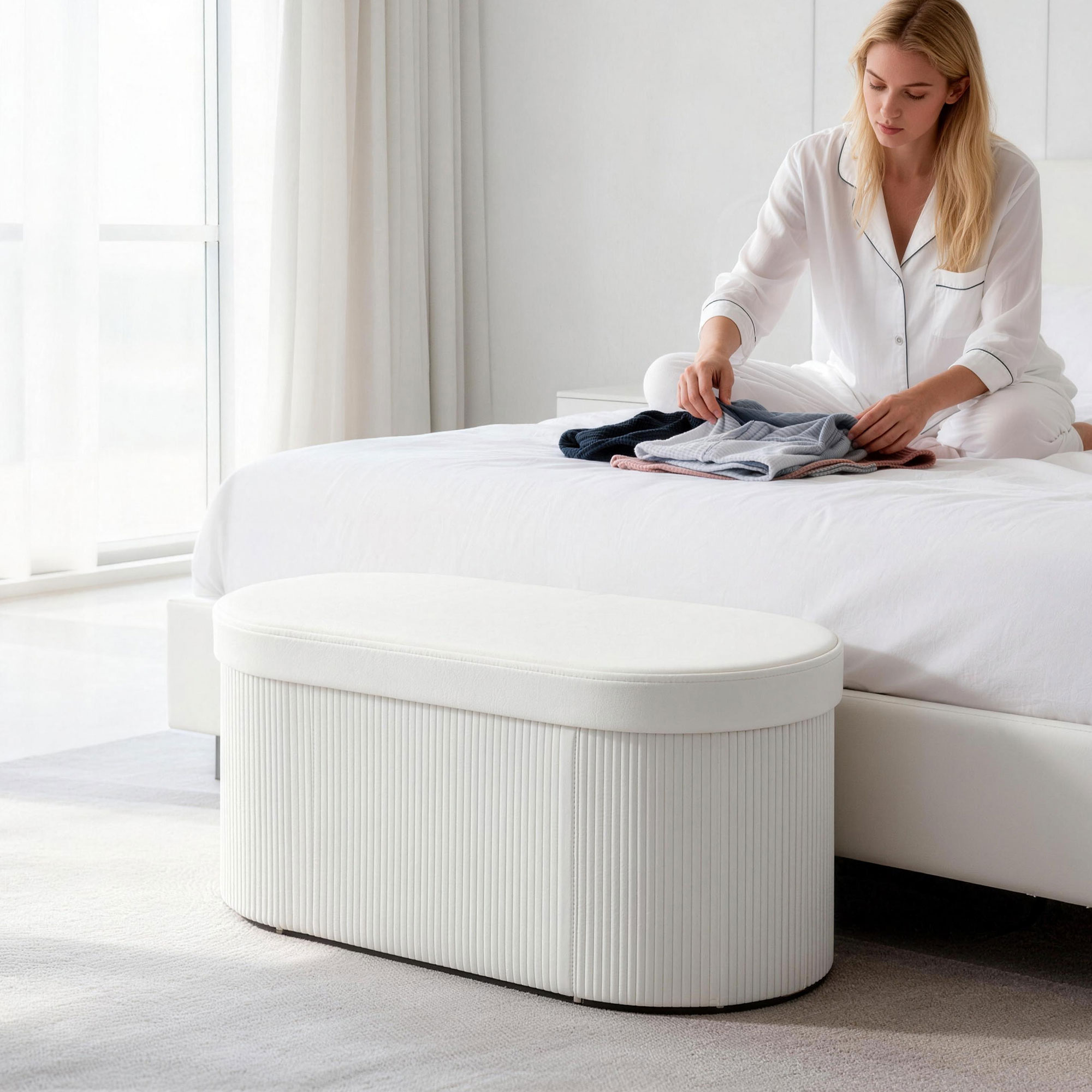 Una mujer dobla ropa en una cama blanca, con un otomano blanco al lado.