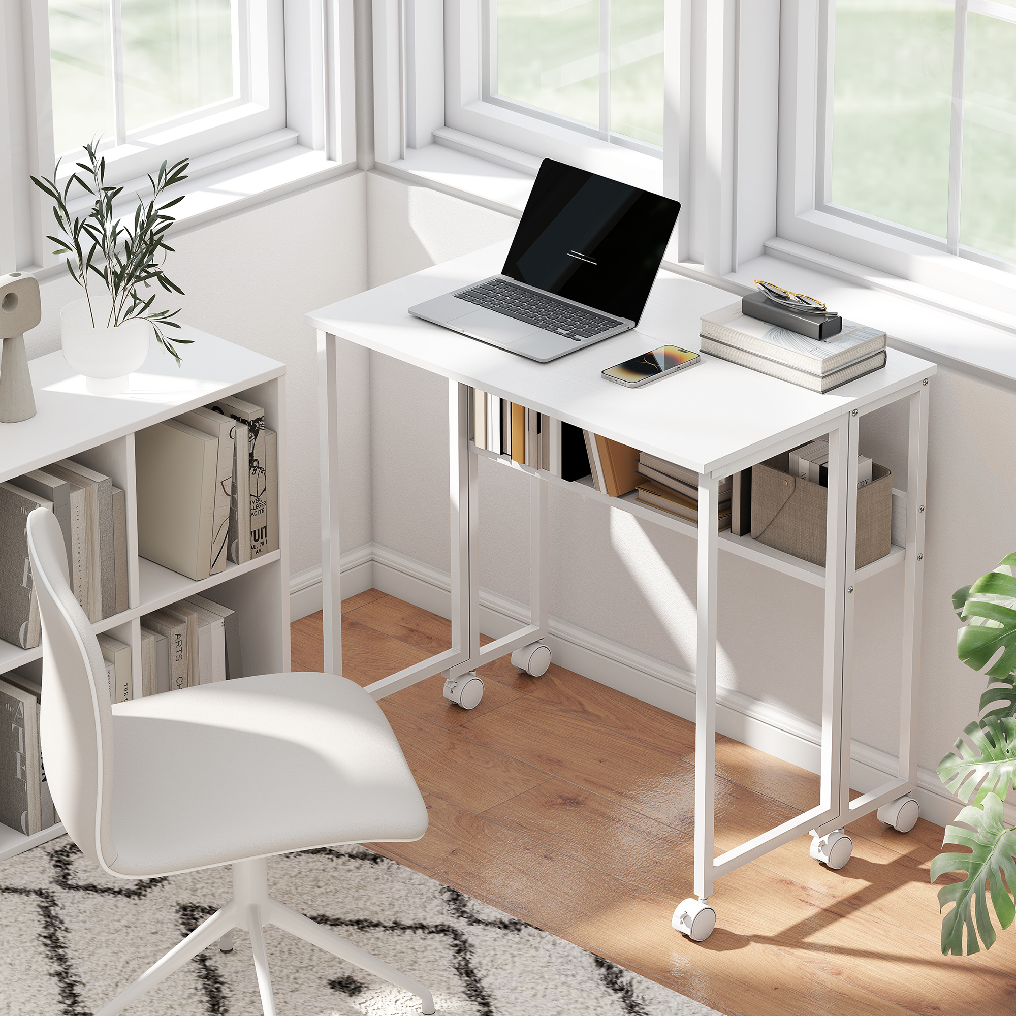 Un escritorio blanco con portátil, teléfono, libros y una estantería al lado.