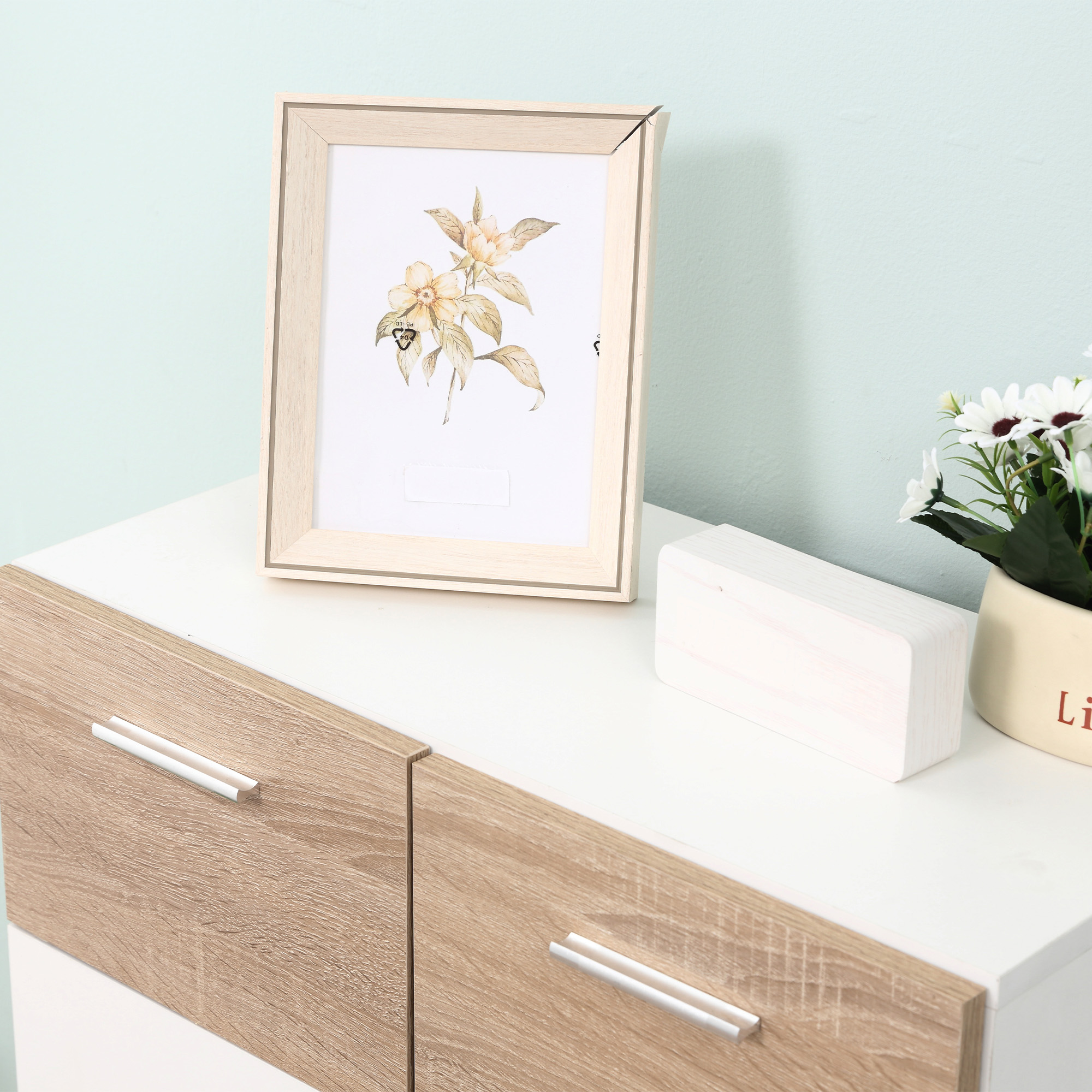 Un marco de fotos de madera clara con flores encima, sobre una cómoda blanca.
