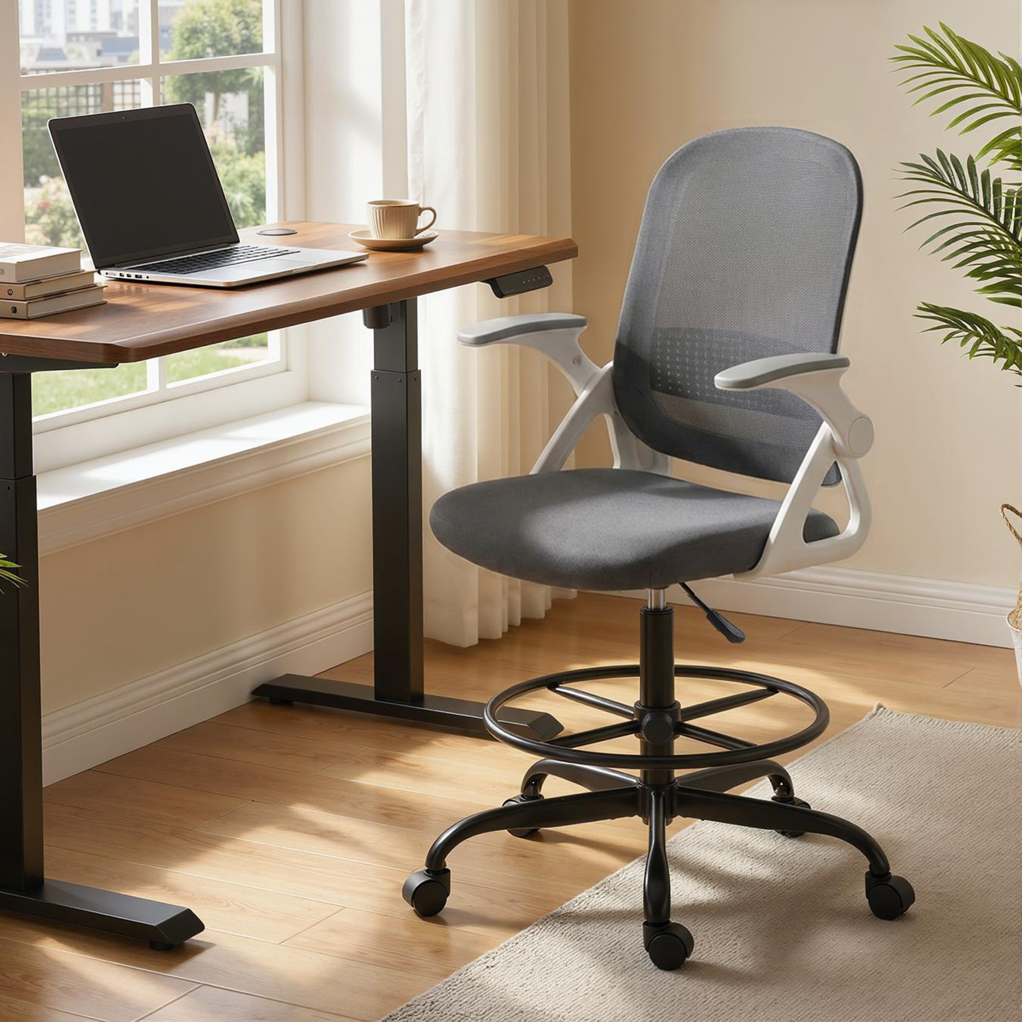 Una silla de malla gris con reposabrazos está al lado de un escritorio ajustable con portátil y taza.