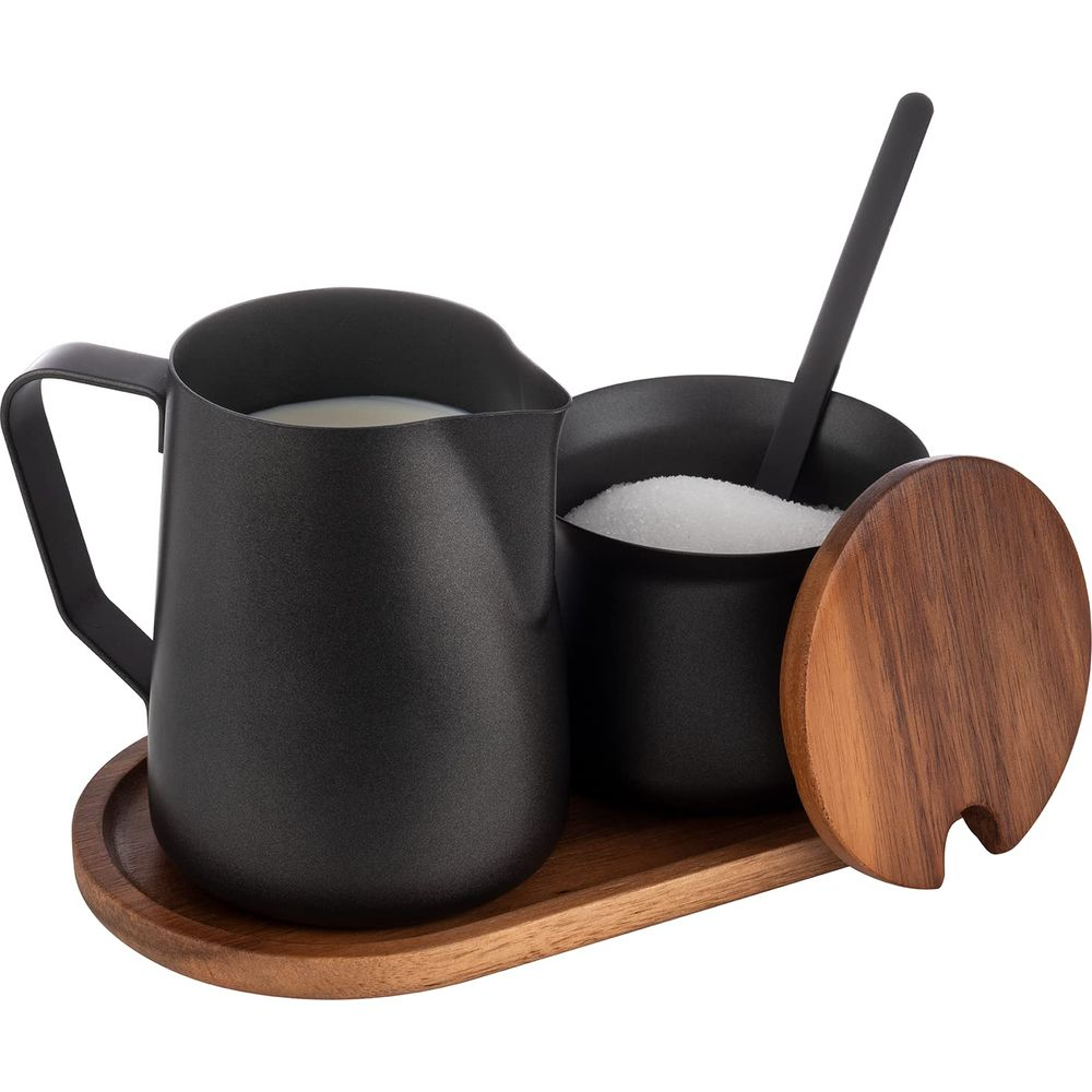 Eine schwarze Kaffeemilchkanne mit Milch und eine Zuckerdose mit Löffel auf einem Holztablett.