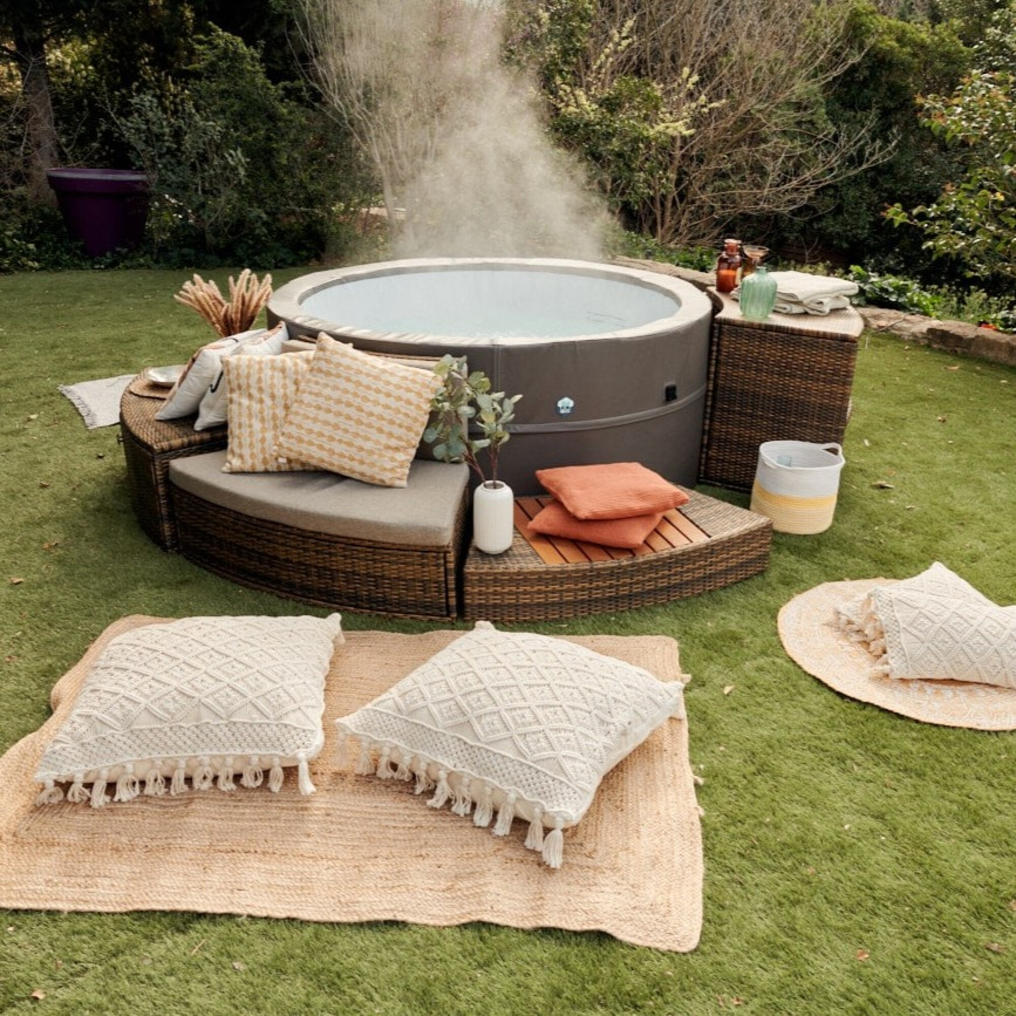 Un jacuzzi inflable sobre césped artificial con cojines y arbustos al fondo.