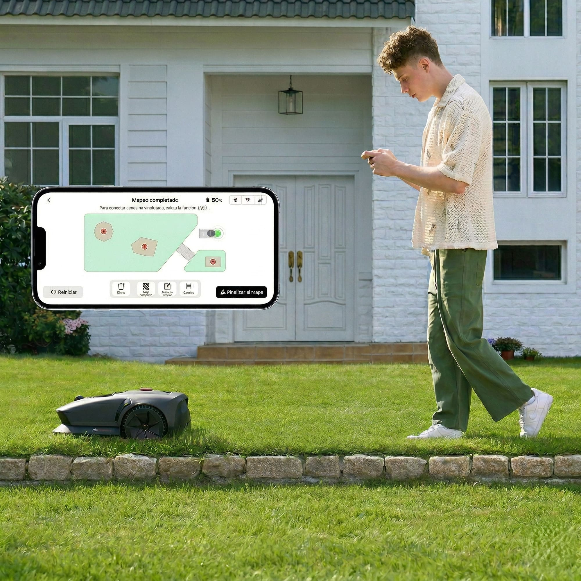 Un joven con teléfono está junto a un robot cortacésped frente a una casa.