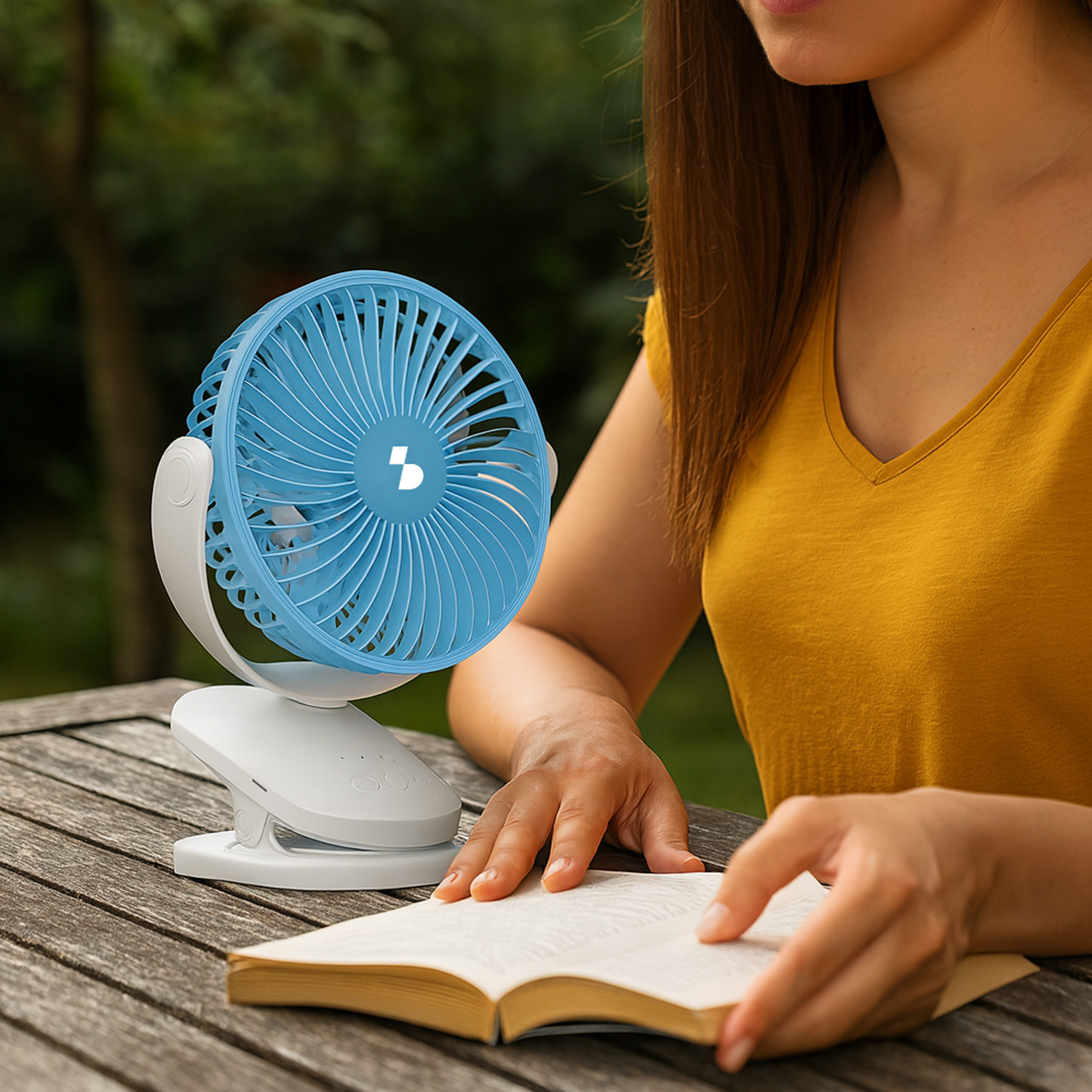 Un ventilatore blu a clip è su un tavolo di legno accanto a una persona che legge un libro.
