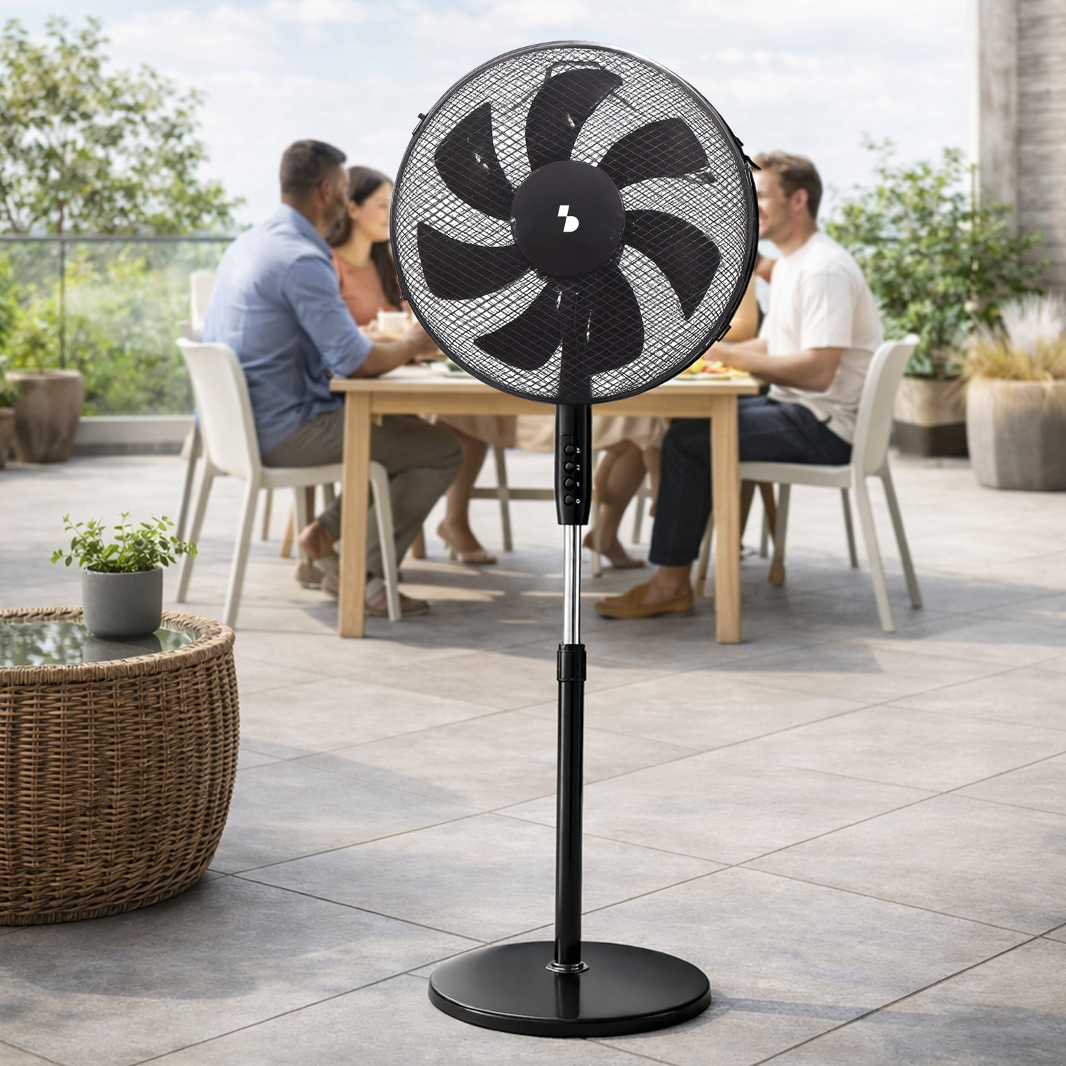 Un ventilatore da terra nero si trova su un patio con persone sedute a un tavolo sullo sfondo.
