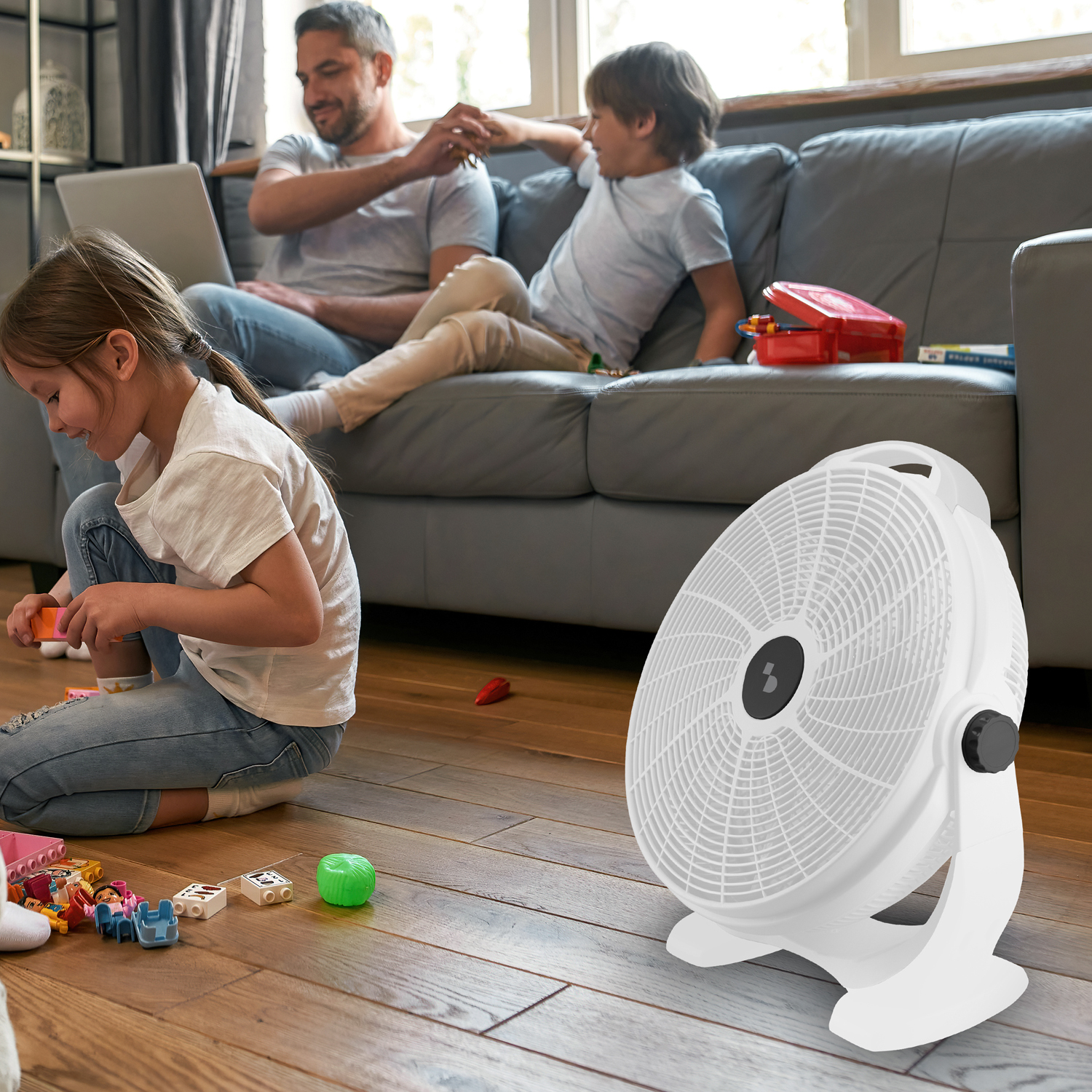 Un ventilatore bianco si trova su un pavimento di legno mentre una famiglia gioca sullo sfondo.