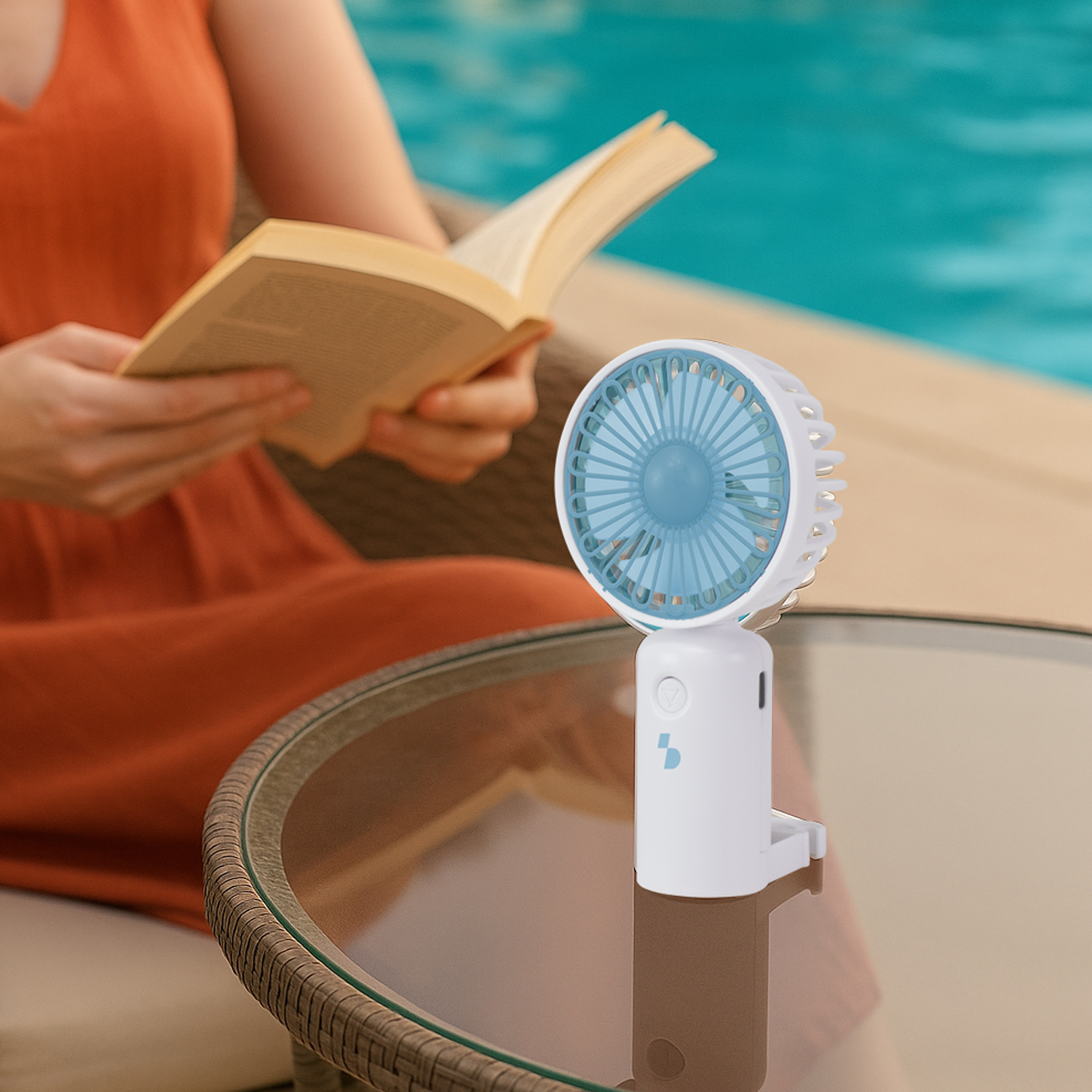 Una persona legge un libro accanto a un piccolo ventilatore su un tavolo vicino a una piscina.