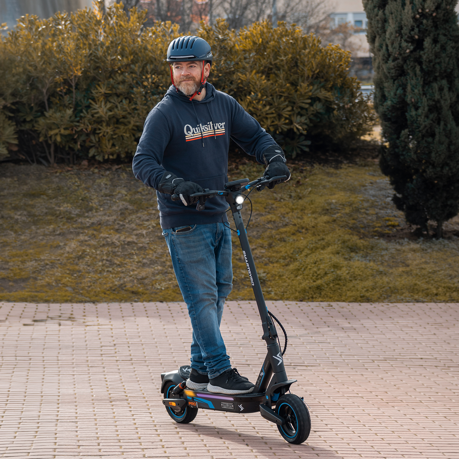 Hombre con casco monta patinete eléctrico por sendero pavimentado, árboles.