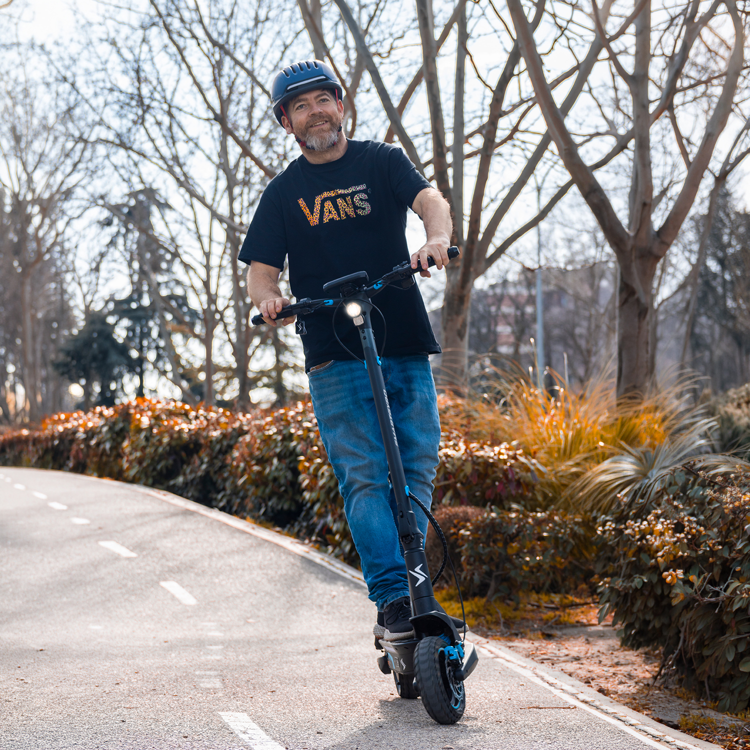 Hombre con camiseta Vans y jeans monta patinete eléctrico en sendero.