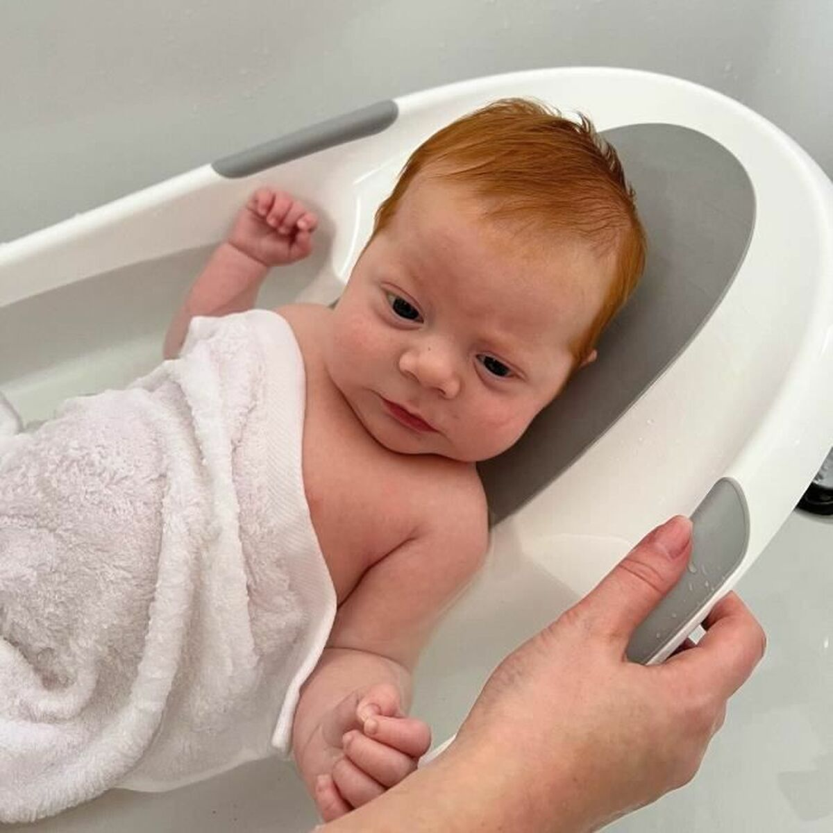 Ein Baby mit roten Haaren liegt in einer Babywanne, eingewickelt in ein weißes Handtuch.