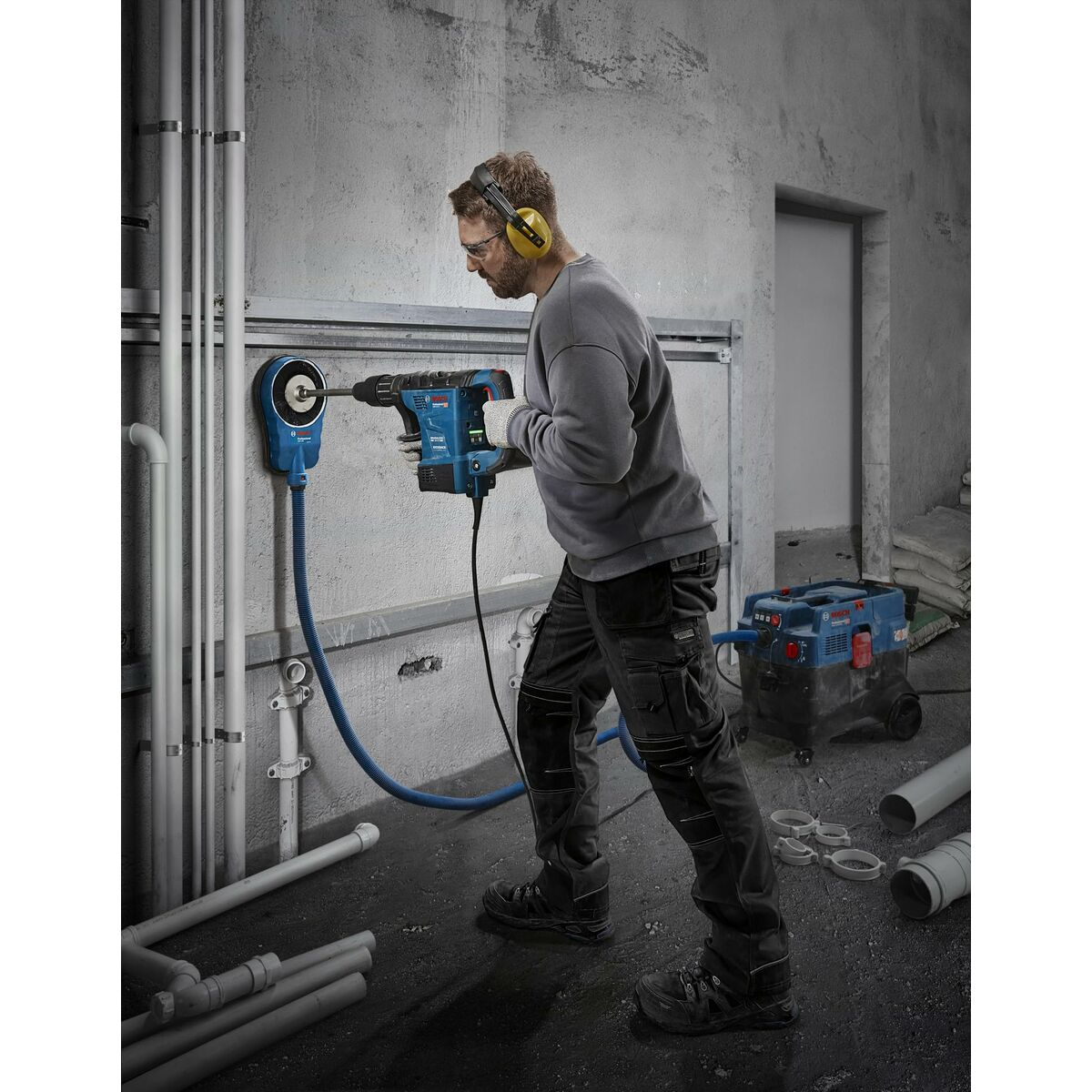 Mann mit Bohrmaschine bohrt in Wand, Staubabsaugung und Rohre.