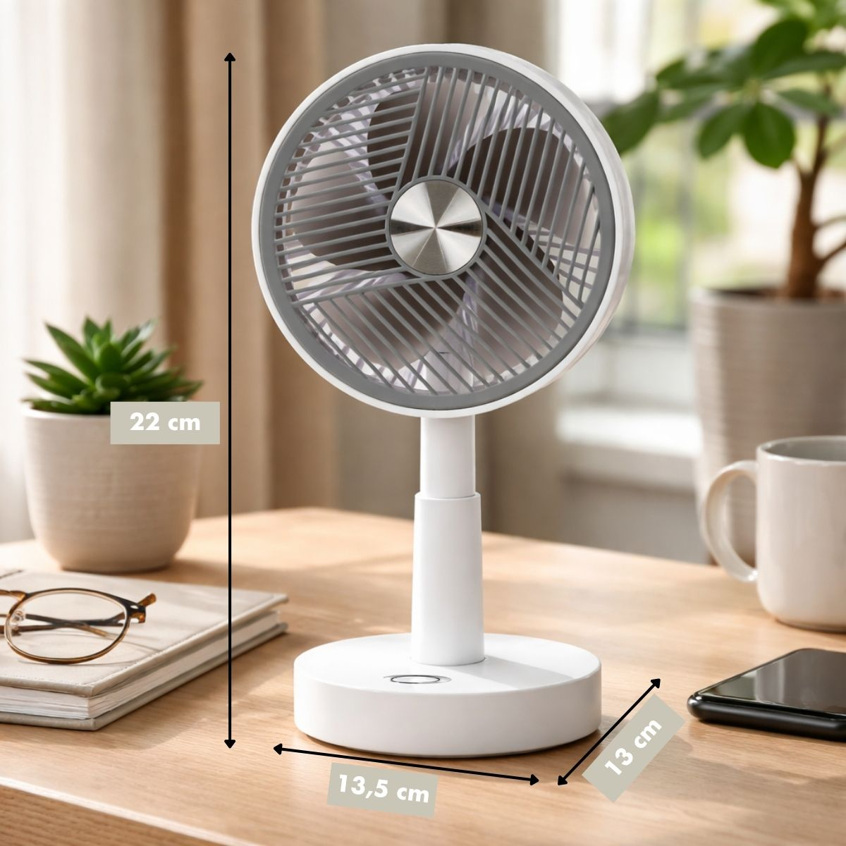 A white desk fan with gray blades sits on a wooden table, with plants and a mug.