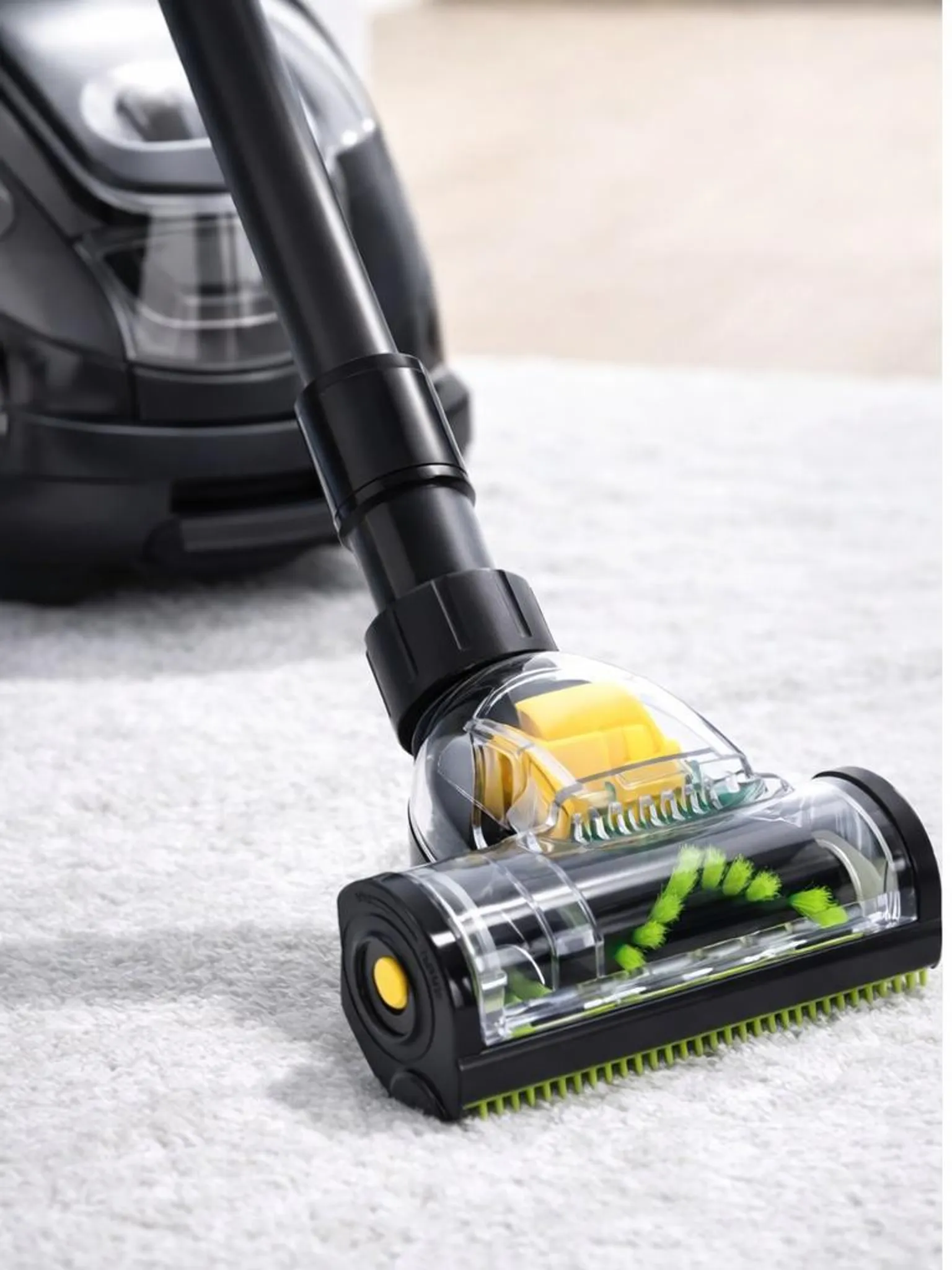 Ein schwarzer Staubsaugerkopf mit transparentem Gehäuse, zeigt gelben Mechanismus und grüne Bürste, auf weißem Teppich.