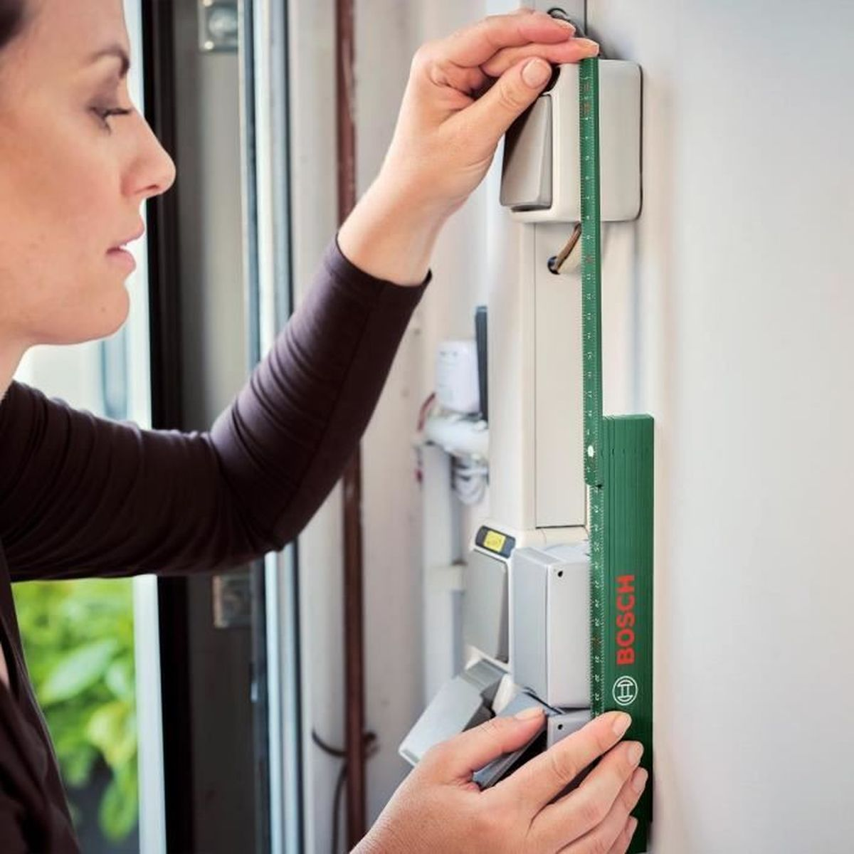 Eine Frau misst mit einem grünen Bosch-Lineal die Höhe an einer Wand mit elektrischen Komponenten.