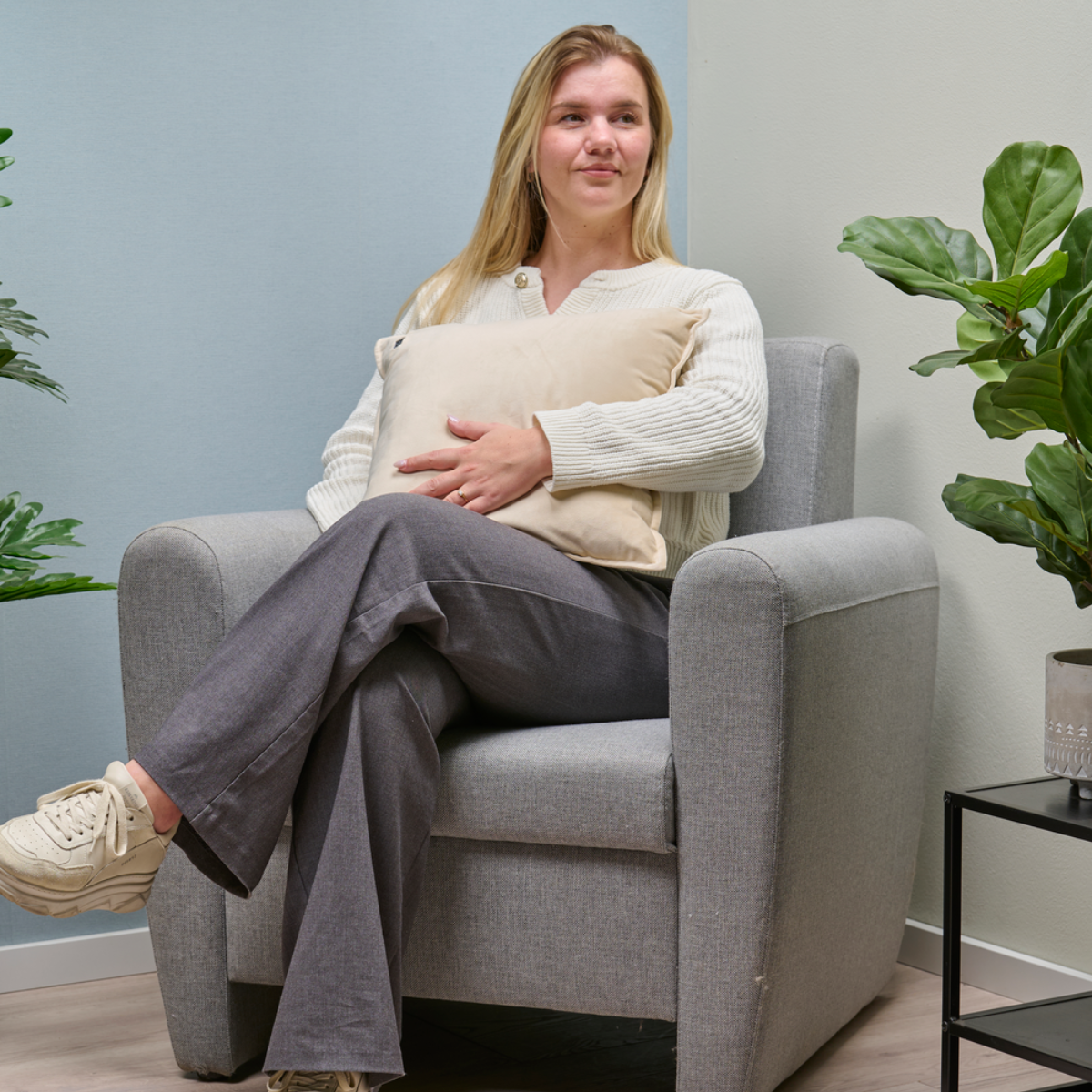 A young woman sits in a grey armchair holding a cream-colored cushion.