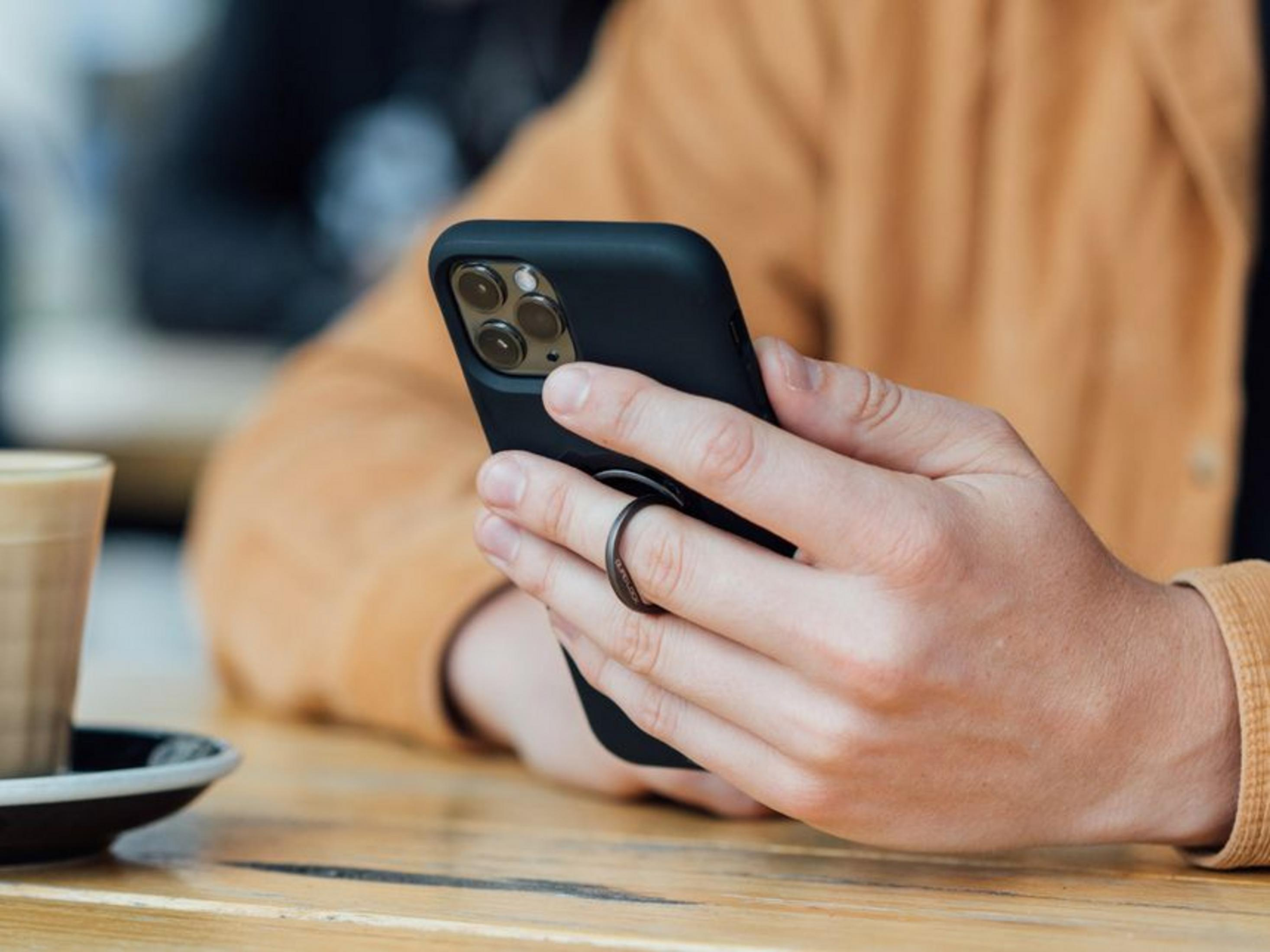 Eine Person hält ein schwarzes Smartphone mit einem Ringhalter und trinkt Kaffee.