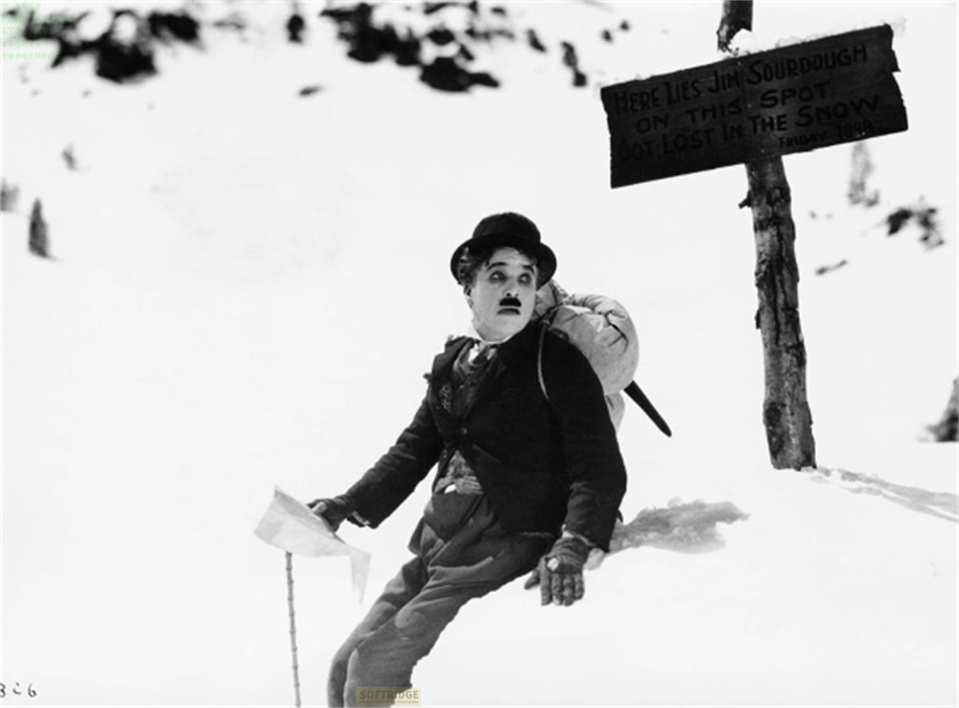 Charlie Chaplin sulla neve con uno zaino, vicino a un cartello.