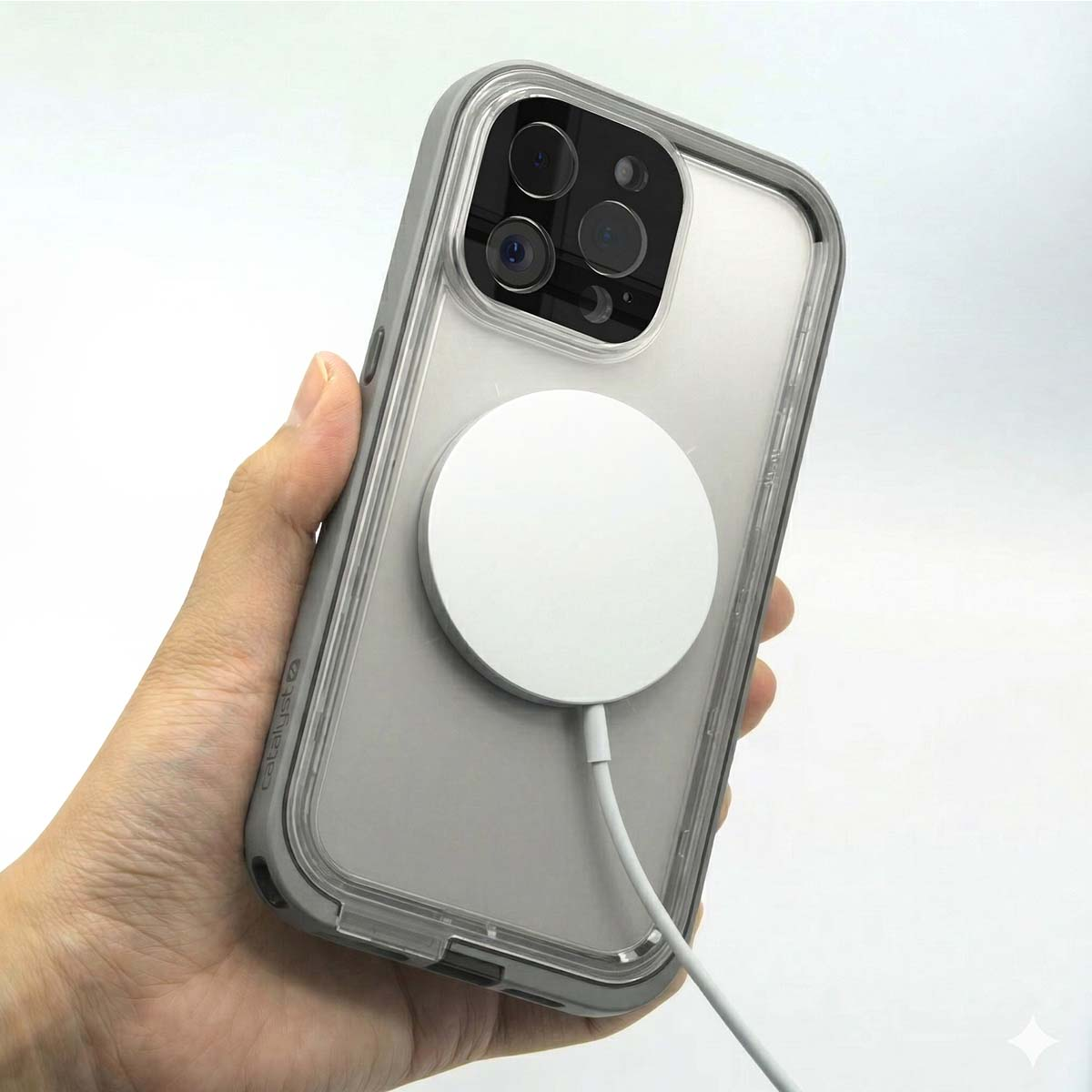Une main tient une coque d'iPhone transparente avec un pare-chocs gris, attachée à un chargeur MagSafe blanc.