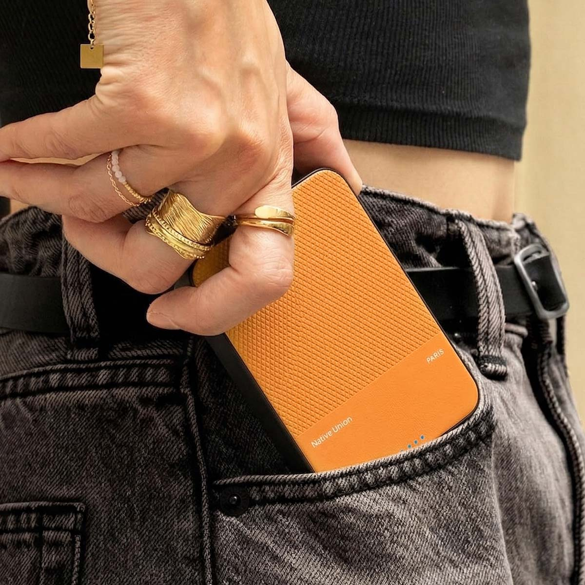 Eine Hand mit Ringen steckt ein orangefarbenes Ladegerät in eine Jeans-Tasche.